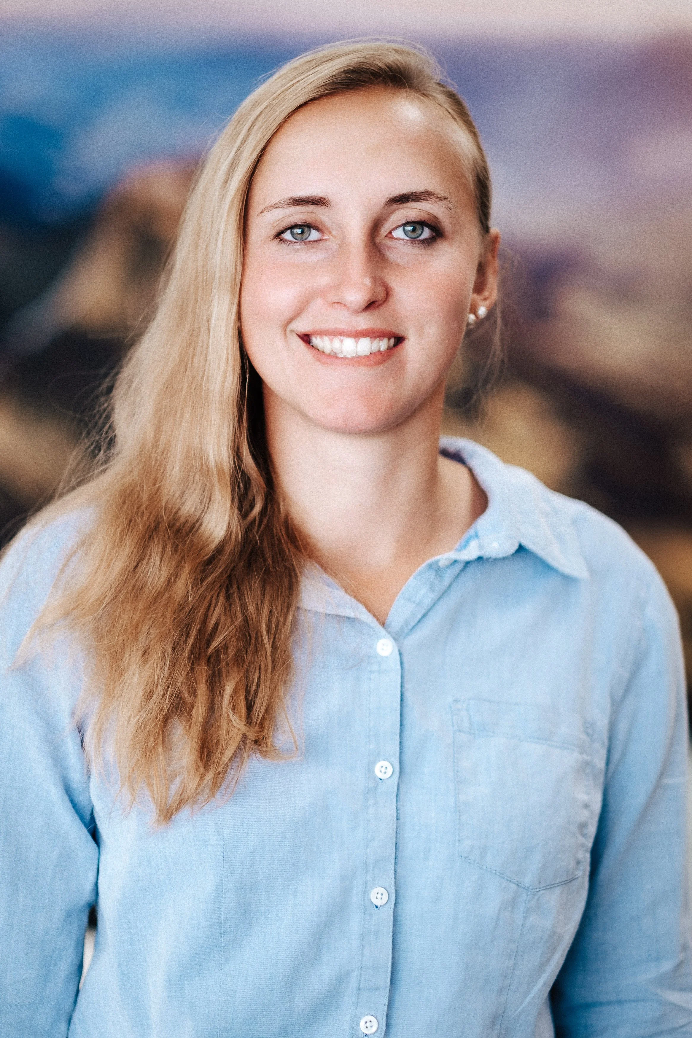 Ein Porträt einer lächelnden Frau mit langen, blonden Haaren, die ein hellblaues Hemd trägt, vor einem unscharfen, natürlichen Hintergrund.