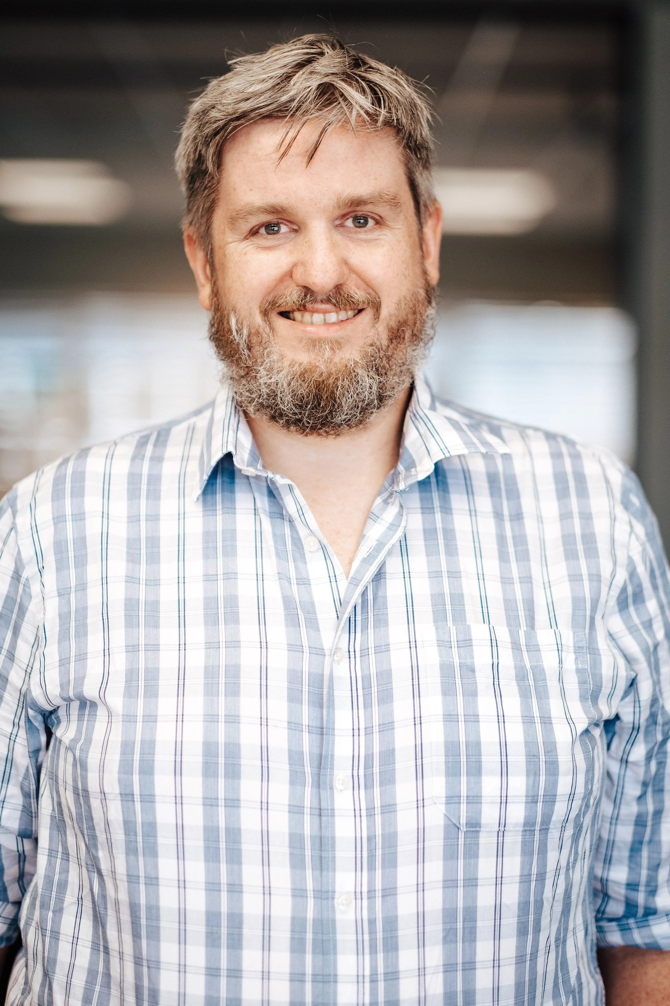 Ein lächelnder Mann mit grauem Bart und kurzen, bräunlichen Haaren, trägt ein blau-weiß gestreiftes Hemd, vor einem modernen, unscharfen Hintergrund.