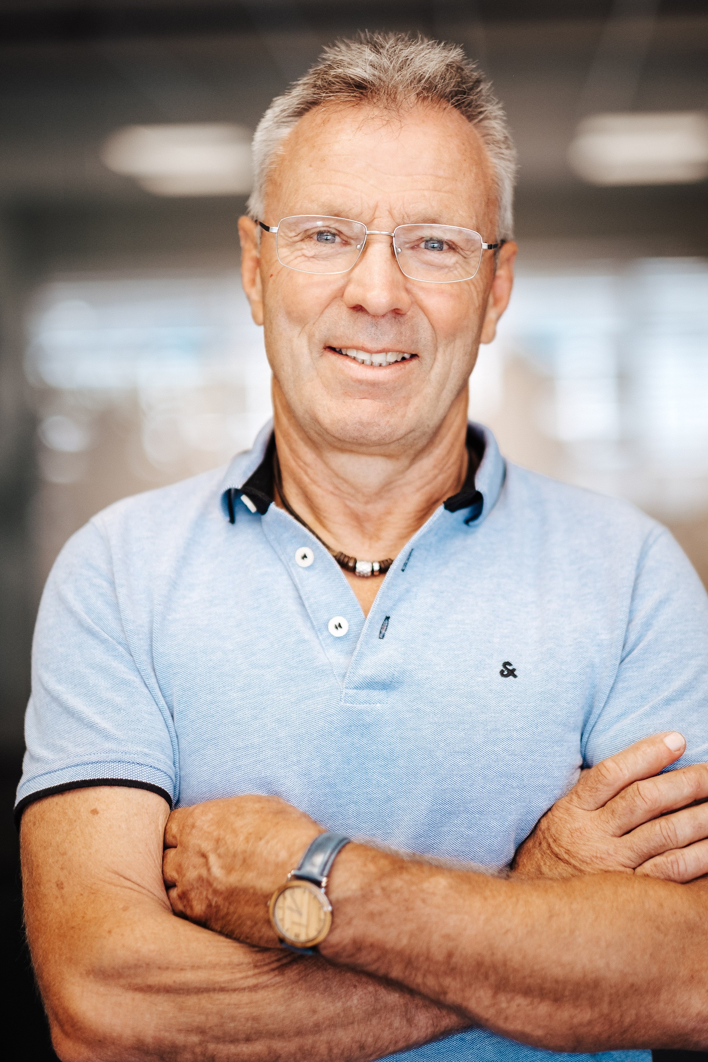 Ein älterer Mann mit Brille und grauem Haar, trägt ein hellblaues Poloshirt, steht mit verschränkten Armen in einem Indoor-Umfeld und lächelt.