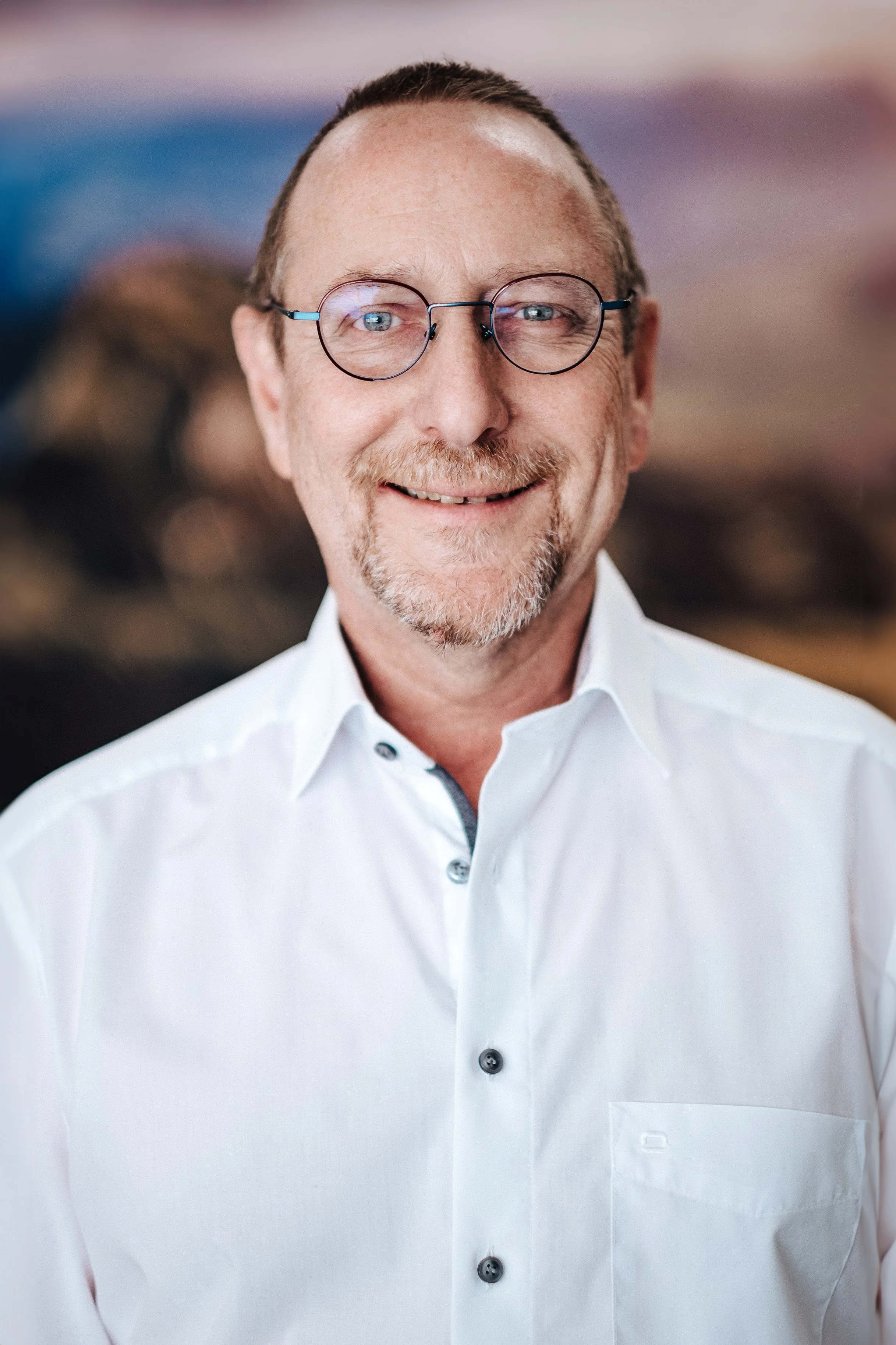 Porträt eines lächelnden Mannes mit Brille in einem weißen Hemd vor einem unscharfen Hintergrund.