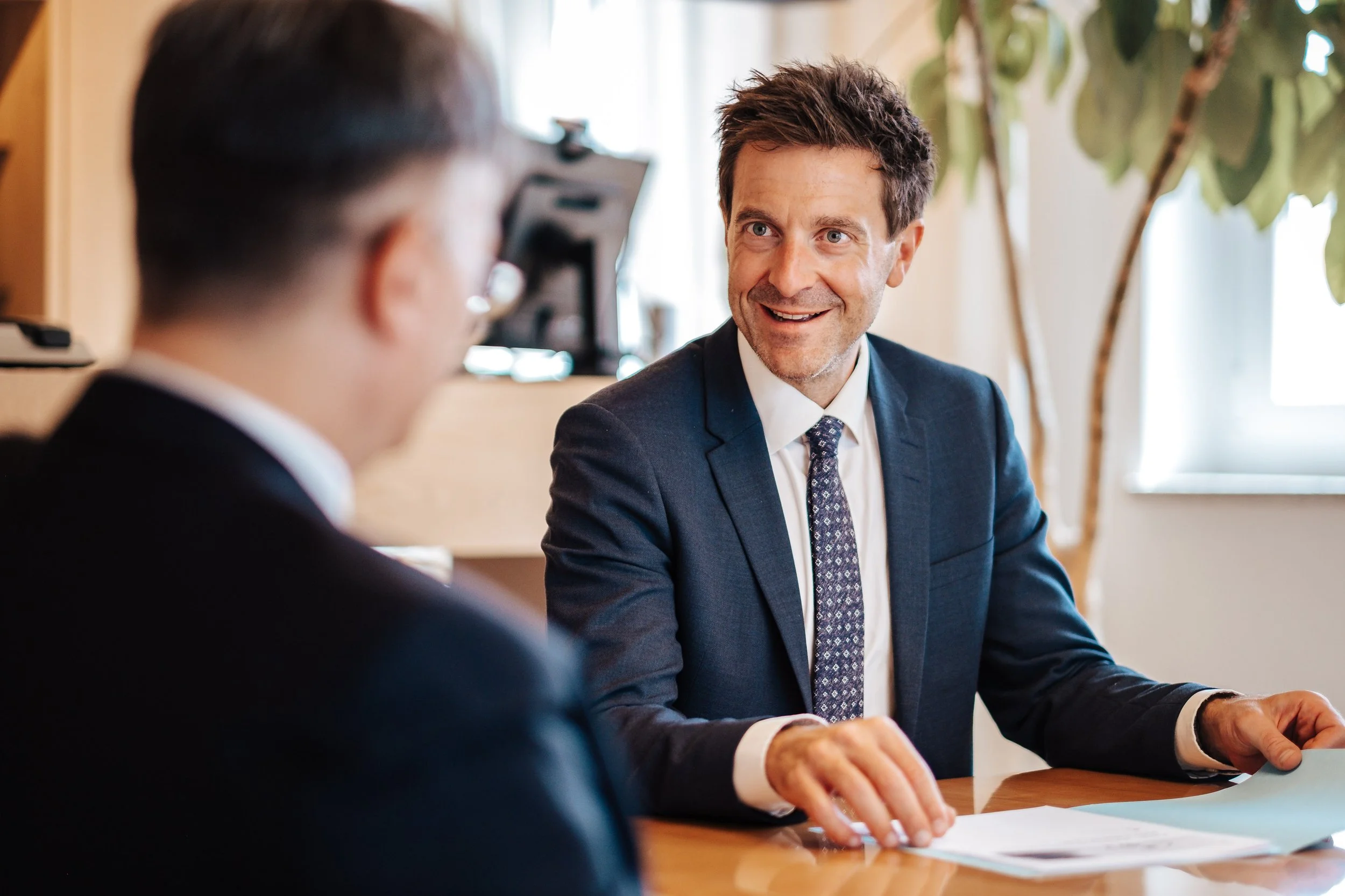 Zwei Männer im Gespräch, einer lächelnd, im Büro mit natürlichem Licht und Pflanzen im Hintergrund.