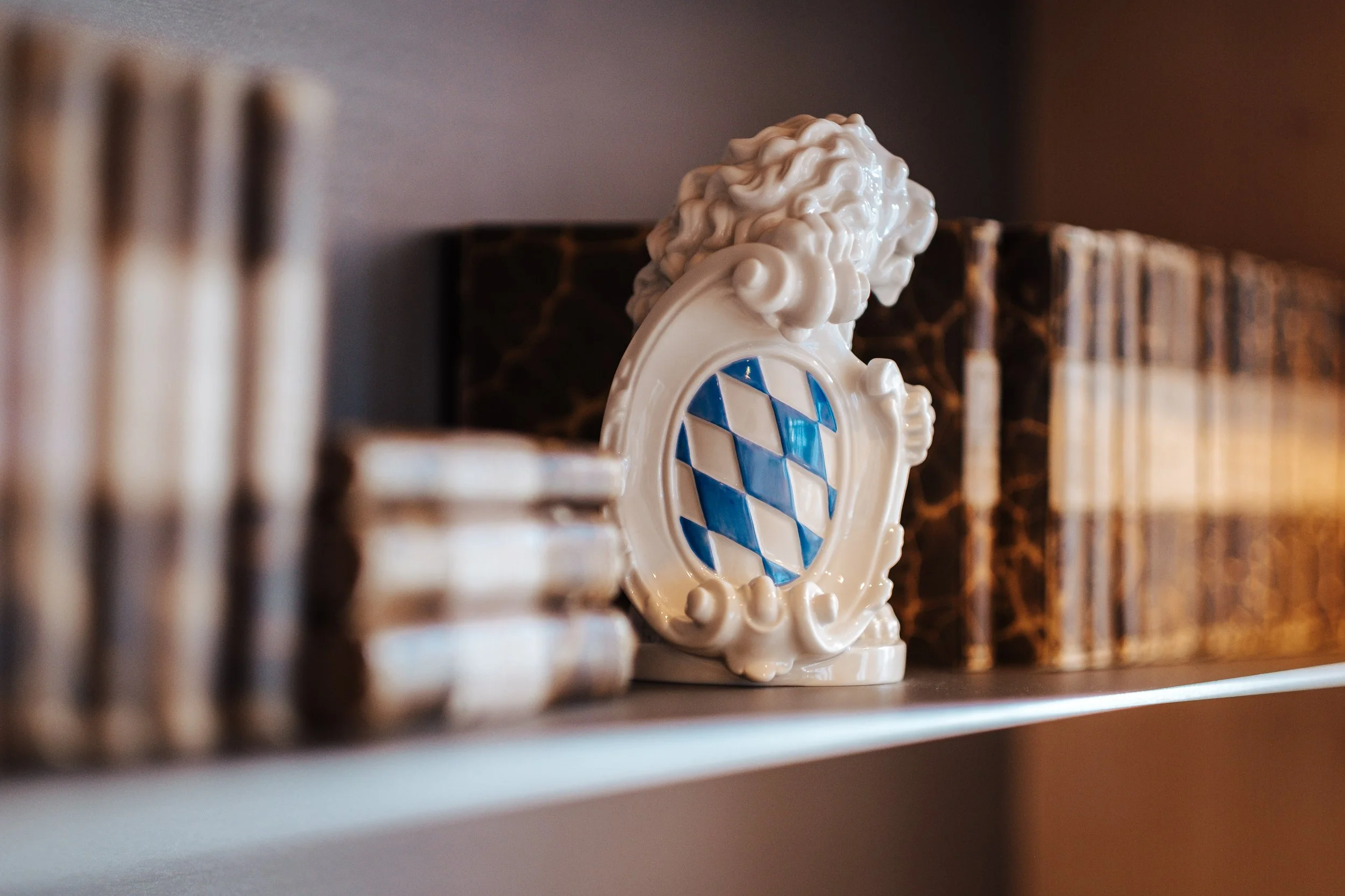 Porzellanfigur mit bayerischem Wappen, Blumentapete und Bücher im Hintergrund.