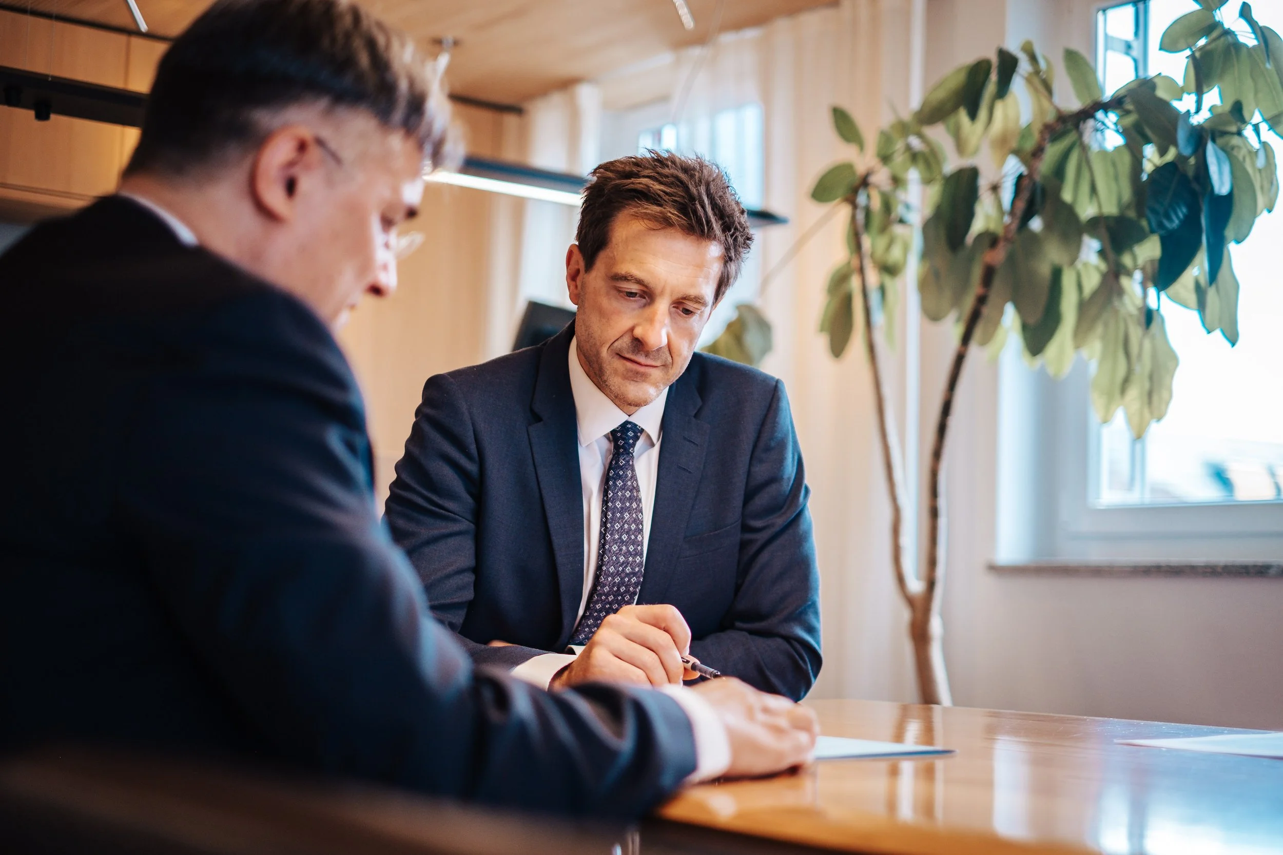 Zwei Männer in Anzügen während eines Meetings in einem Büro, mit einem großen Fenster im Hintergrund und einem großen grünen Pflanze.