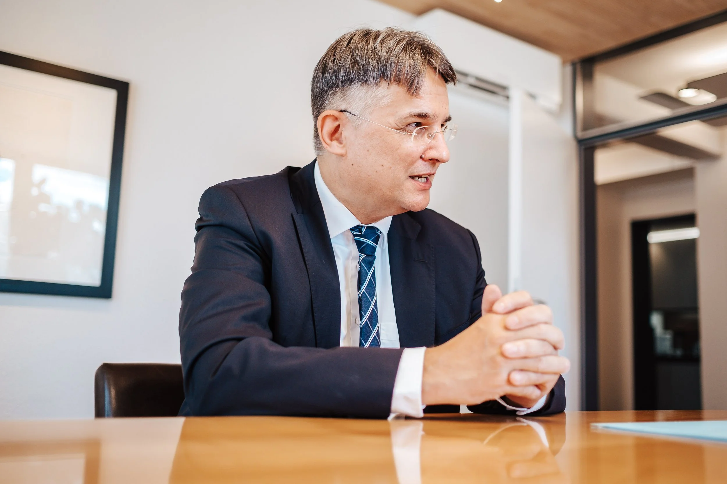 Ein Mann in Anzug sitzt an einem Tisch in einem modernen Büro, er scheint im Gespräch zu sein.