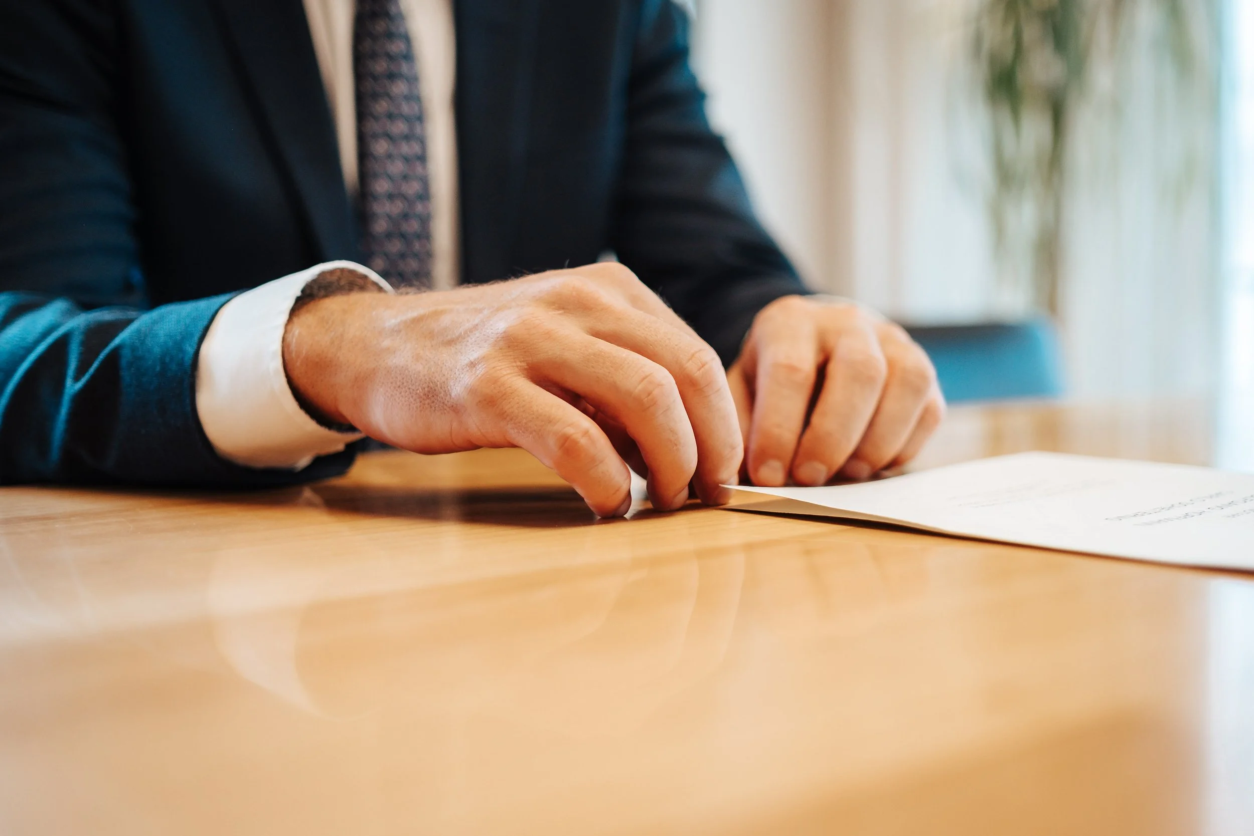 Ein Mann in einem dunklen Anzug und einer karierten Krawatte sitzt an einem Holztisch, schreibt auf ein Papier.