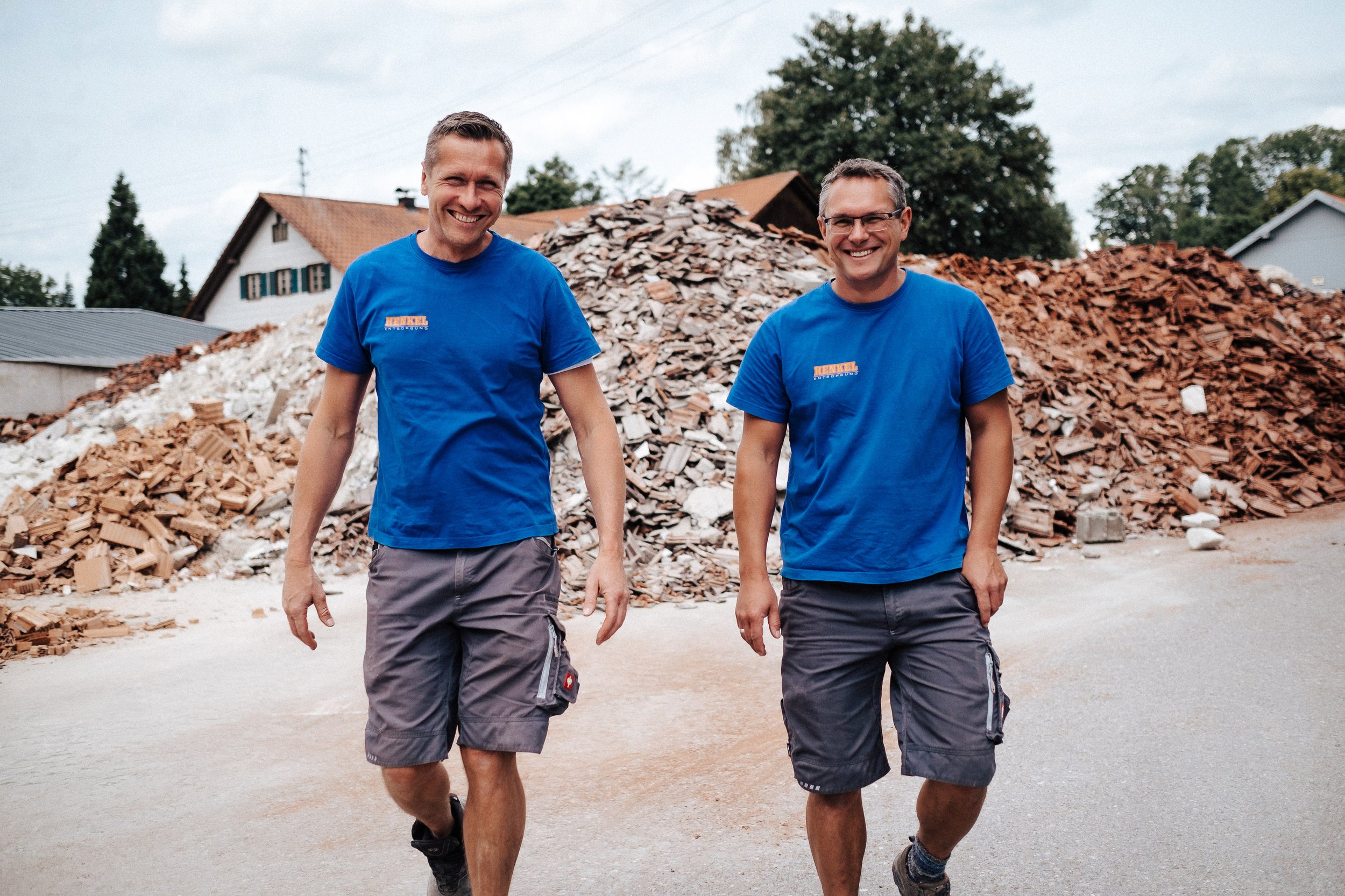 Zwei Männer in blauen T-Shirts und Shorts laufen vor einem großen Haufen Schutt und Ziegelsteinen.
