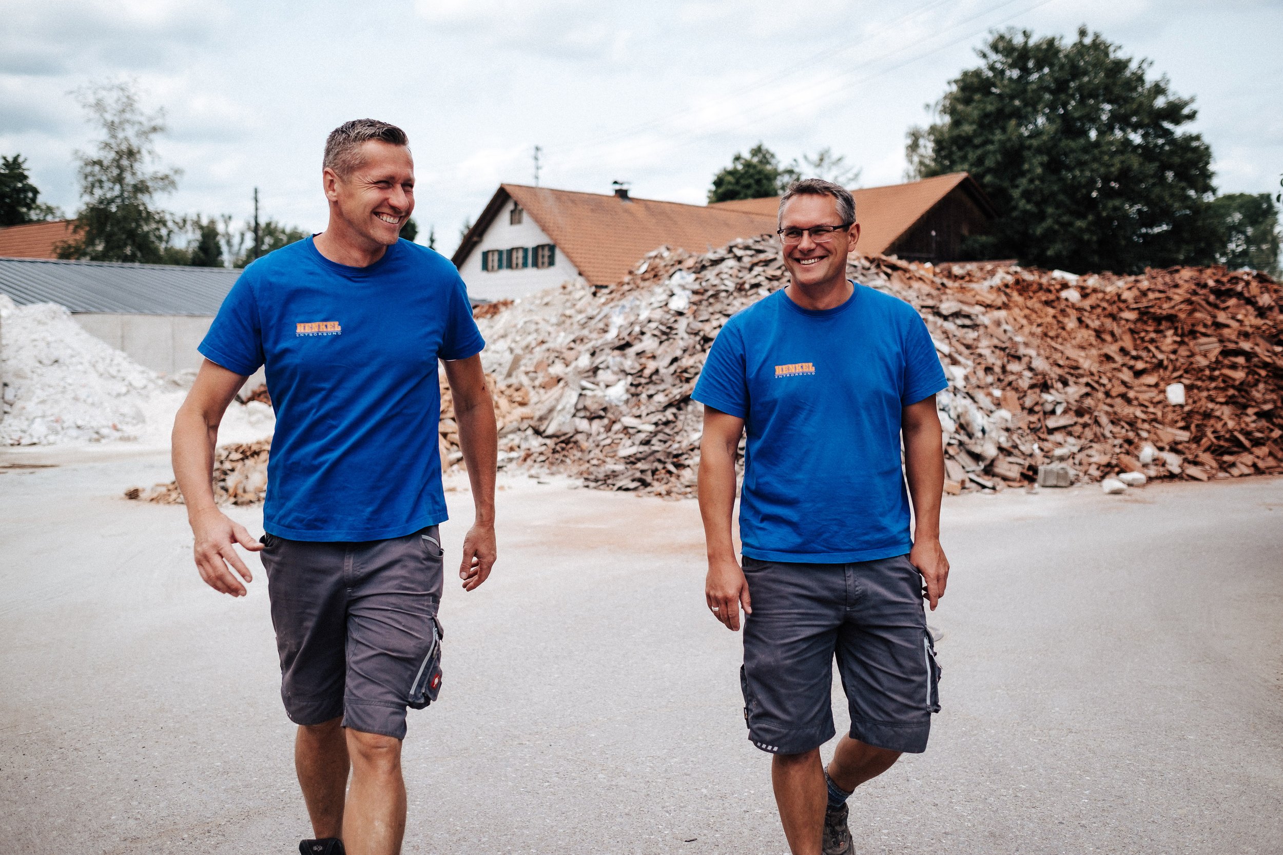 Zwei Männer in blauen T-Shirts und Shorts gehen auf einer Straße mit Laubhaufen im Hintergrund, lachen und unterhalten sich.