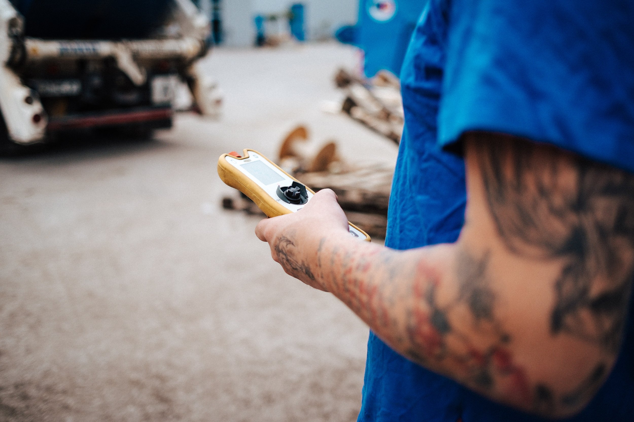Person mit Tattoo am Arm hält ein Messgerät in der Hand, im Hintergrund stehen Baumstämme und ein umgekehrtes Auto.