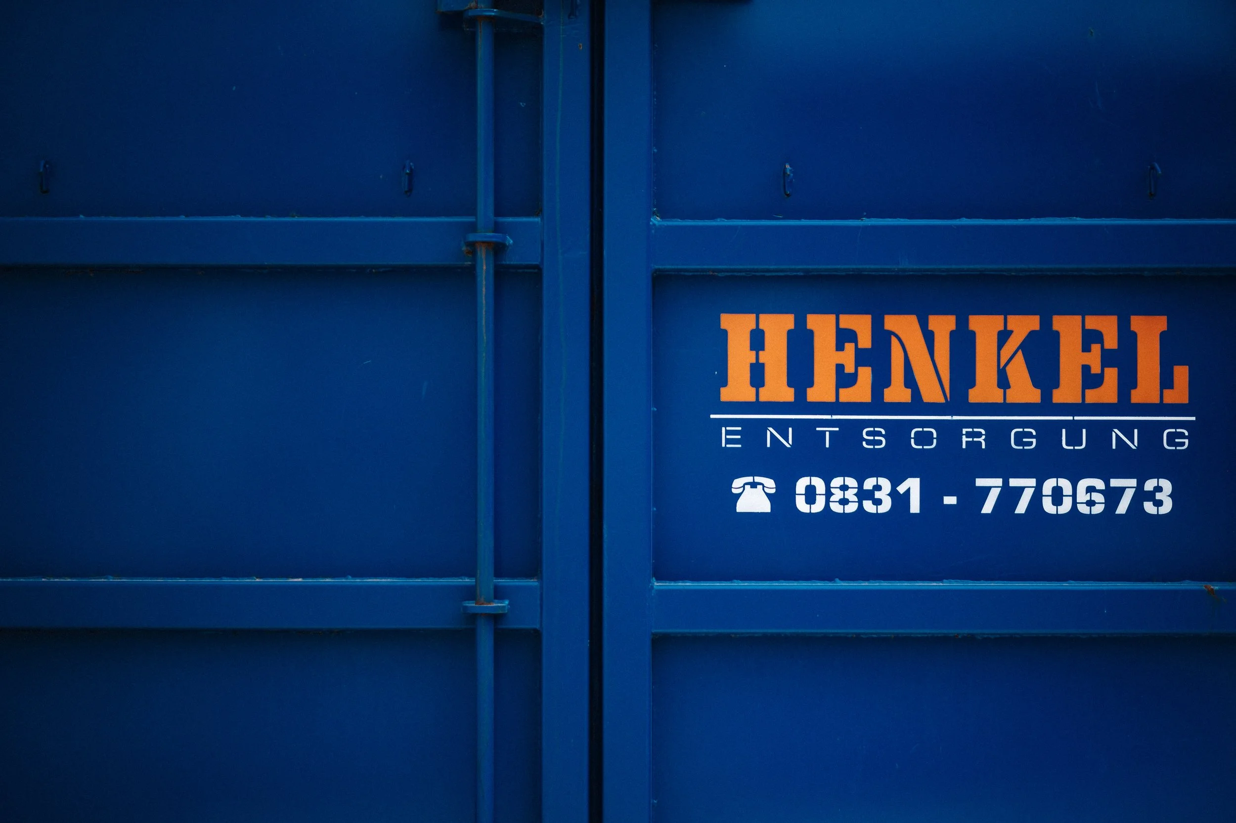 Blaue Metalltür mit der Aufschrift 'HENKEL Entsorgung' in oranger Farbe, darunter ein Telefonhörer-Symbol und die Telefonnummer 0831-770673.