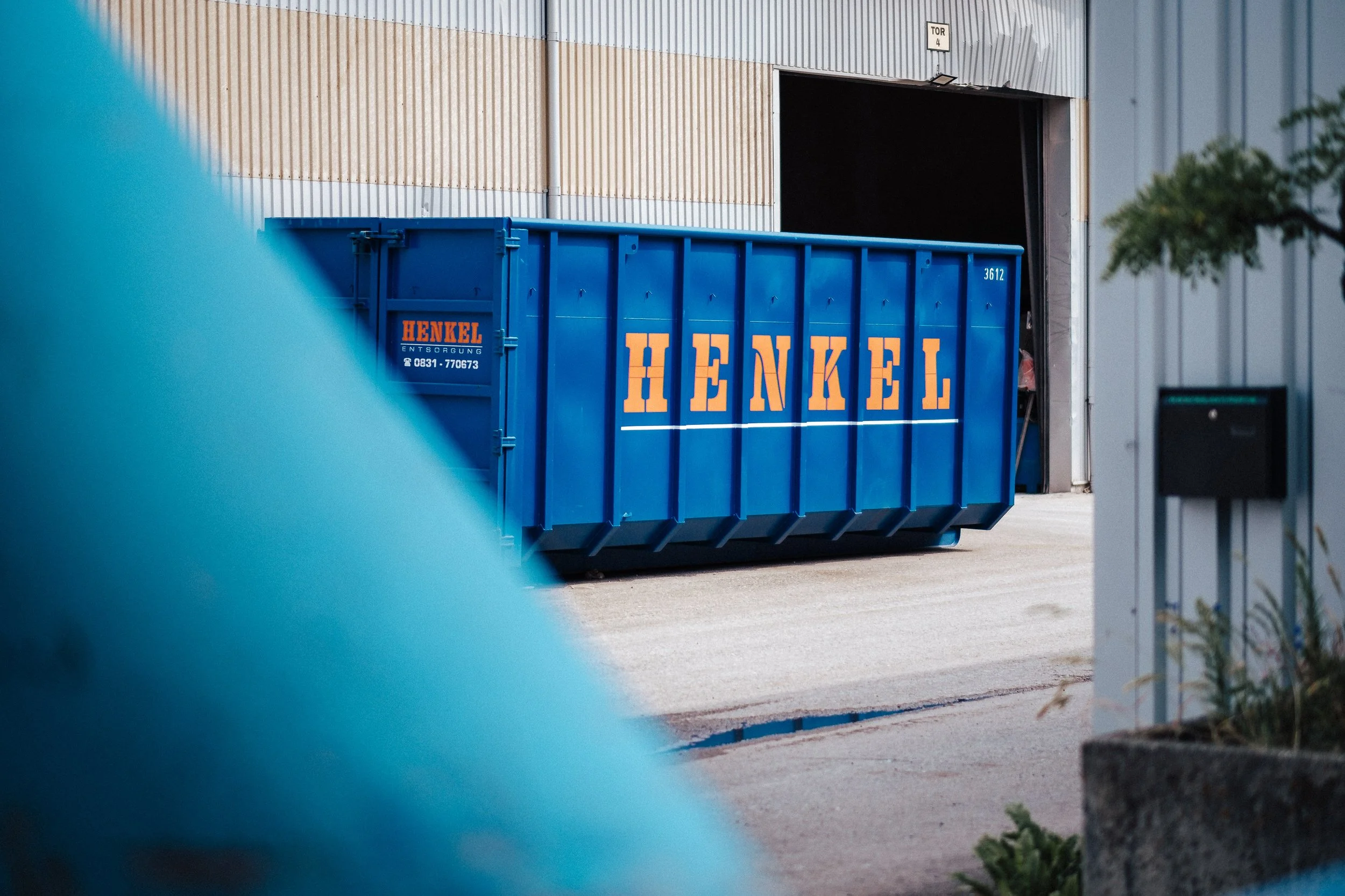 Ein blauer Müllcontainer mit dem Schriftzug 'HENKEL' steht vor einer Industriehalle.