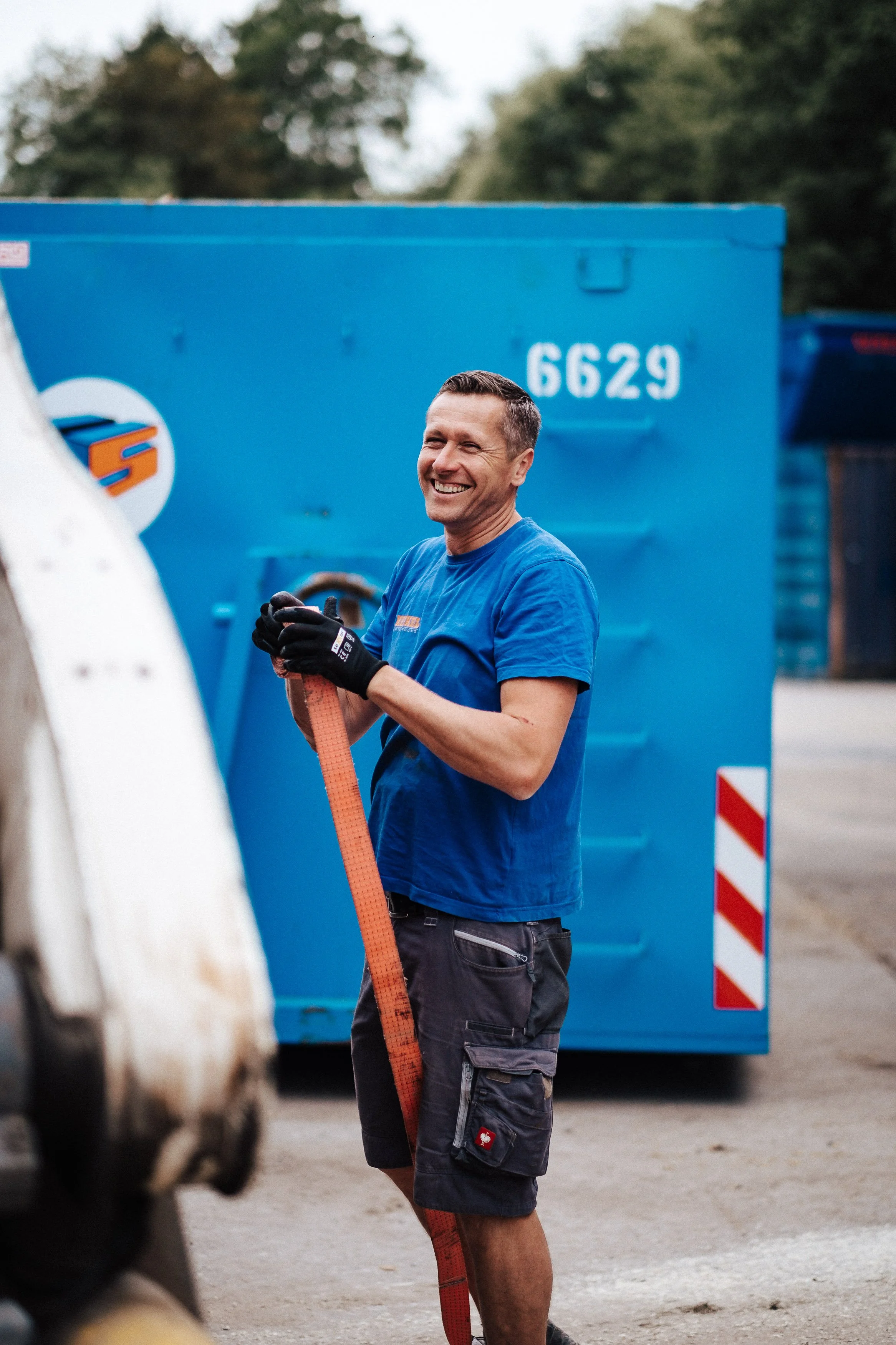 Ein Mann trägt blaue Arbeitskleidung und hält ein rotes Sicherheitsband, steht vor einem blauen Container, lächelt und wirkt fröhlich.