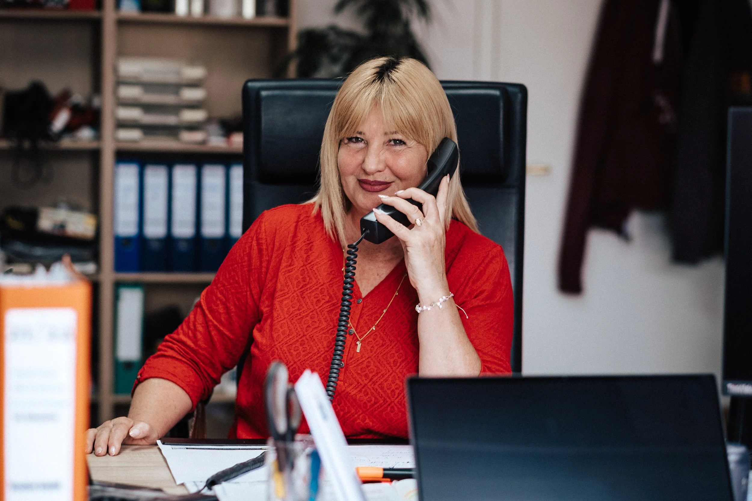 Frau mit blonden Haaren in rotem Hemd, die im Büro am Schreibtisch sitzt und telefoniert.
