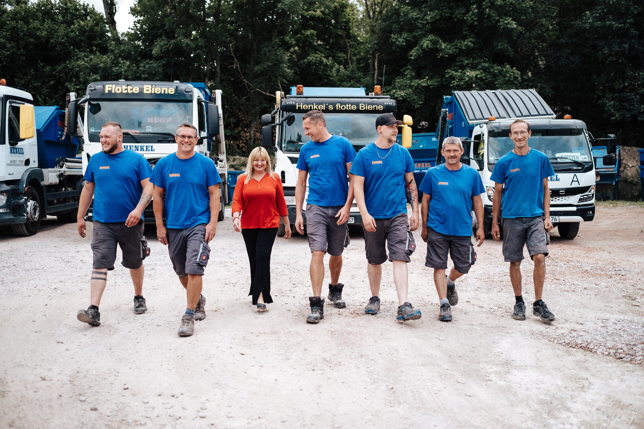 Eine Gruppe von sechs Menschen, fünf Männer und eine Frau, läuft auf einem Bauplatz mit Baustellenfahrzeugen im Hintergrund, lächelnd und in Arbeitshosen.
