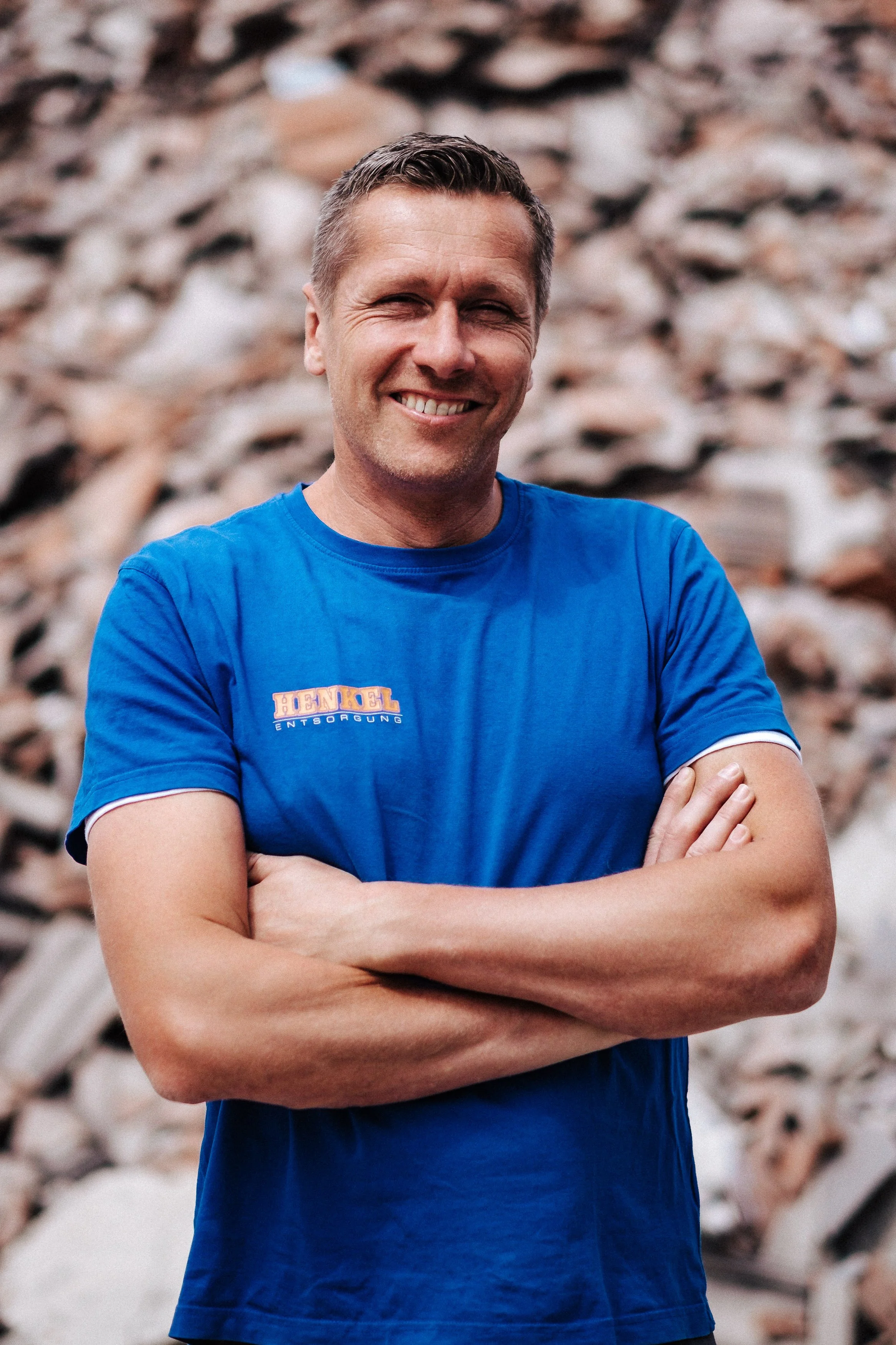 Ein lachender Mann in blauer T-Shirt vor einem Holzhaufen.