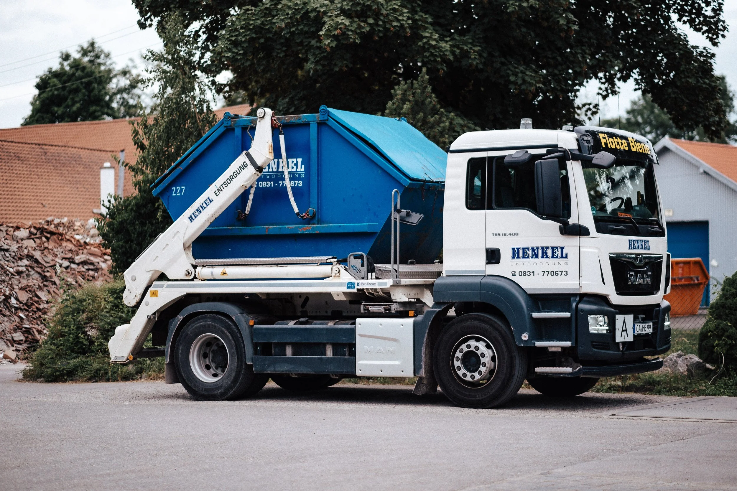 Ein Müllwagen steht am Straßenrand, mit einer blauen Abdeckung auf der Ladefläche, auf der die Aufschrift 'Henkel Entsorgung' zu lesen ist.