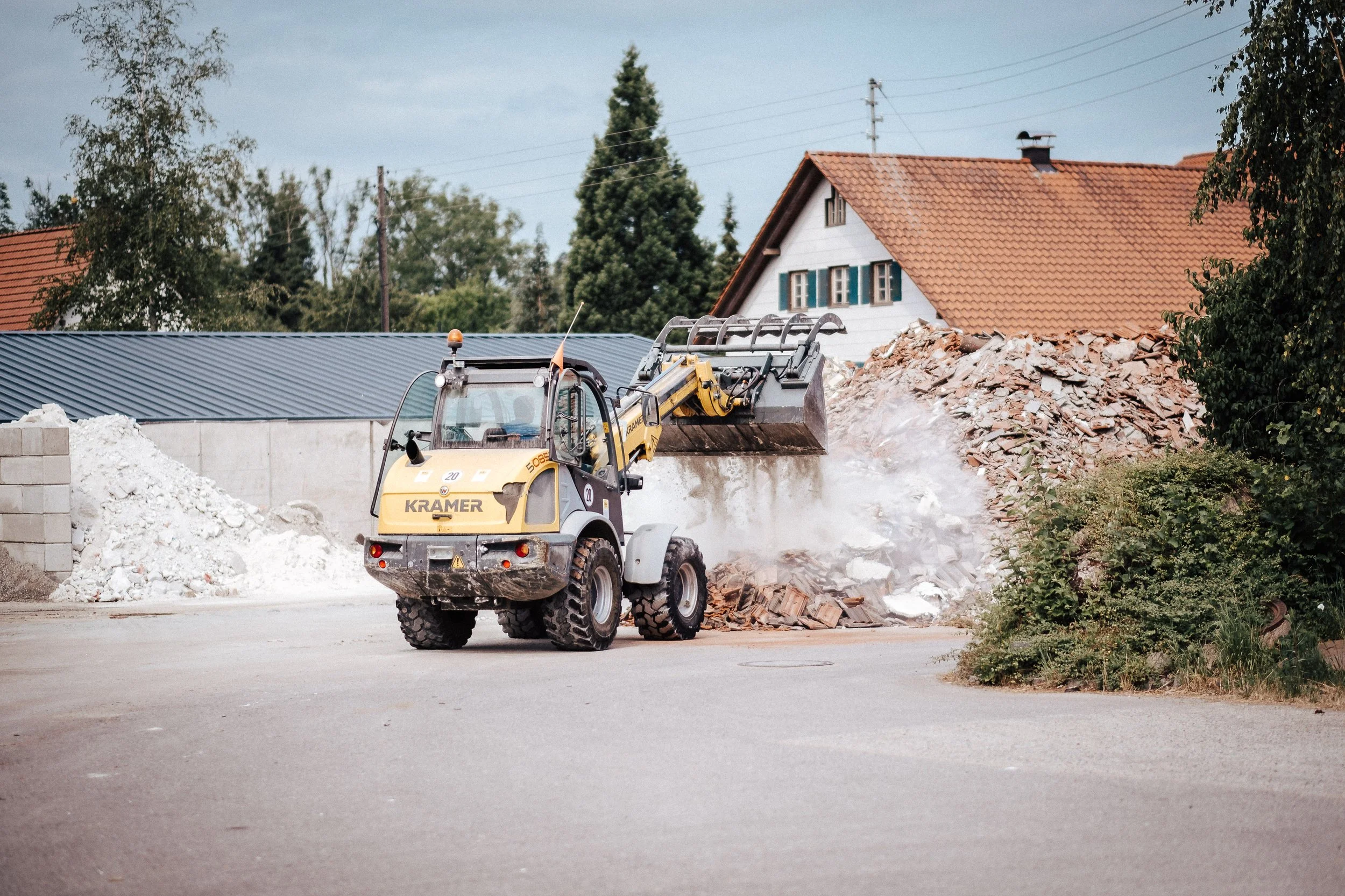 Ein Bagger arbeitet auf einer Baustelle und hebt Bauschutt auf, mit einem Haus mit rot-gelber Ziegeldach im Hintergrund.