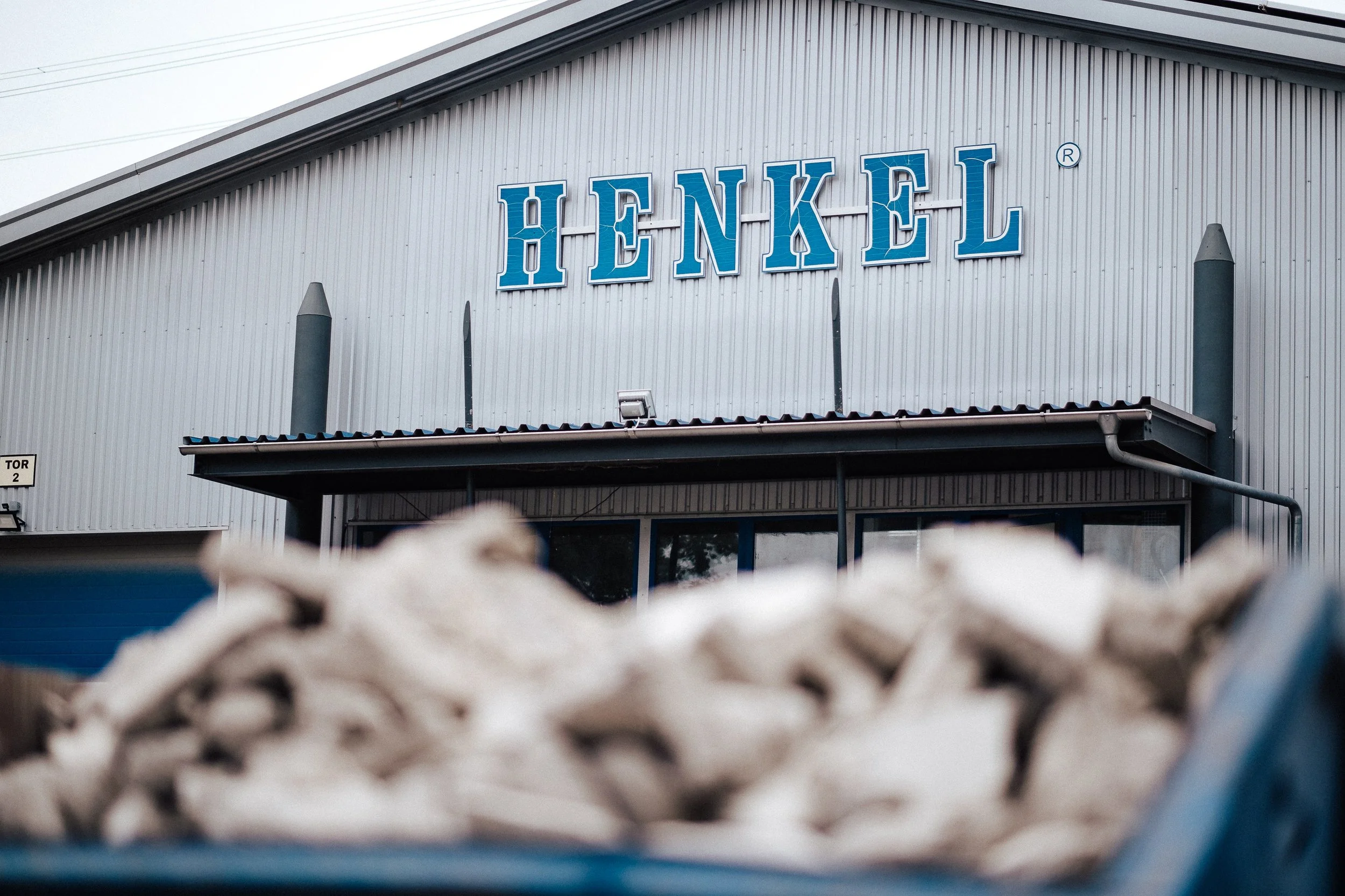 Außenansicht eines Gebäudes mit dem Schriftzug 'HENKEL' an der Fassade, mit einem Container voller zerdrückter Pappkartons im Vordergrund.