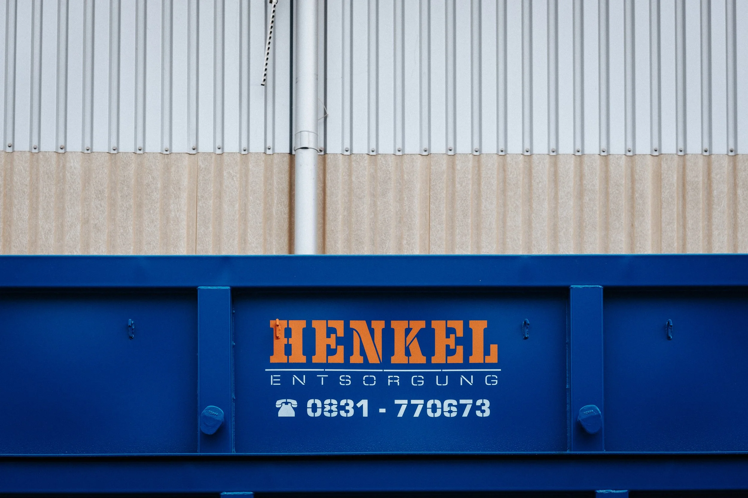 Stahlcontainer mit der Beschriftung 'Henkel Entsorgung' und einer Telefonnummer, vor einer Wand aus Metallplatten und Putz