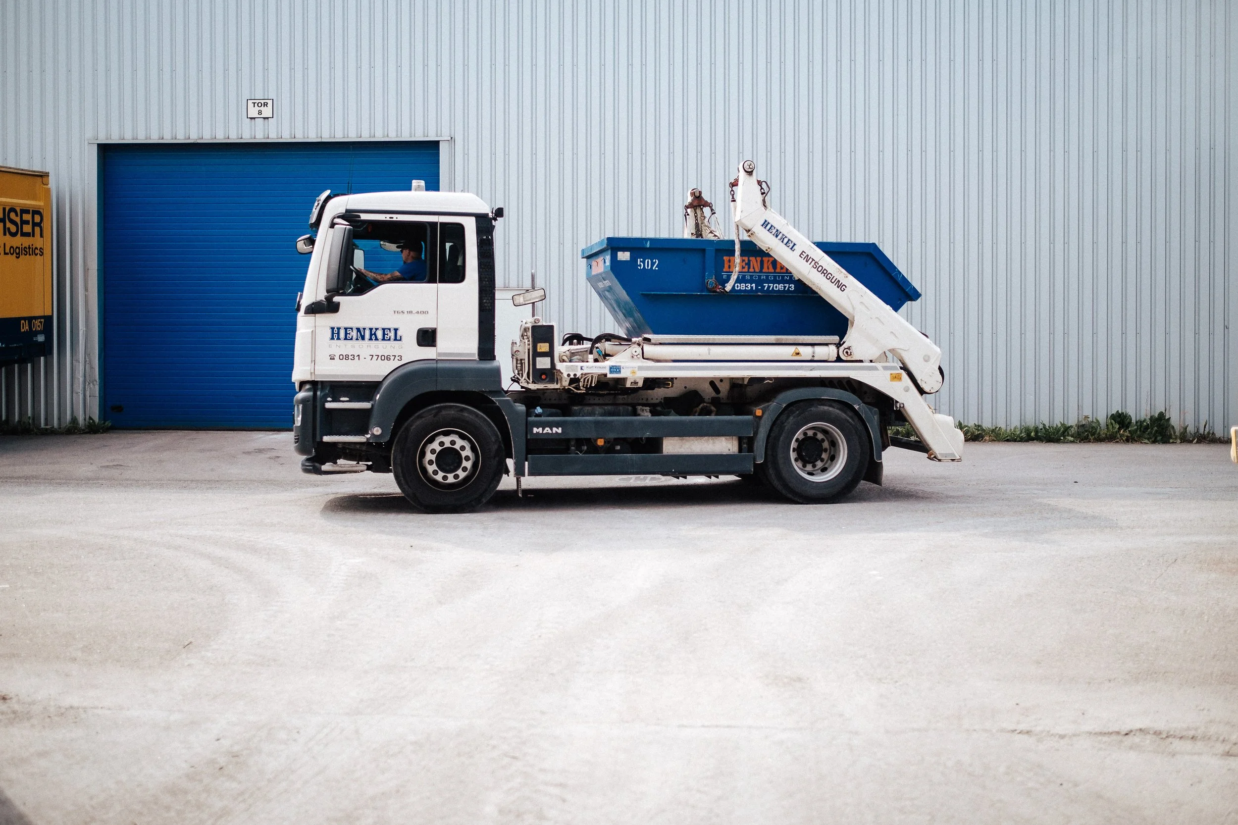 Ein Lieferwagen mit der Beschriftung 'Henkel Entsorgung' steht vor einer grauen Wand mit blauer Türe. Das Fahrzeug ist ein MAN mit einem blauen Container auf der Ladefläche und einem hydraulischen Arm zum Heben. Es wird von einem Fahrer gesteuert.