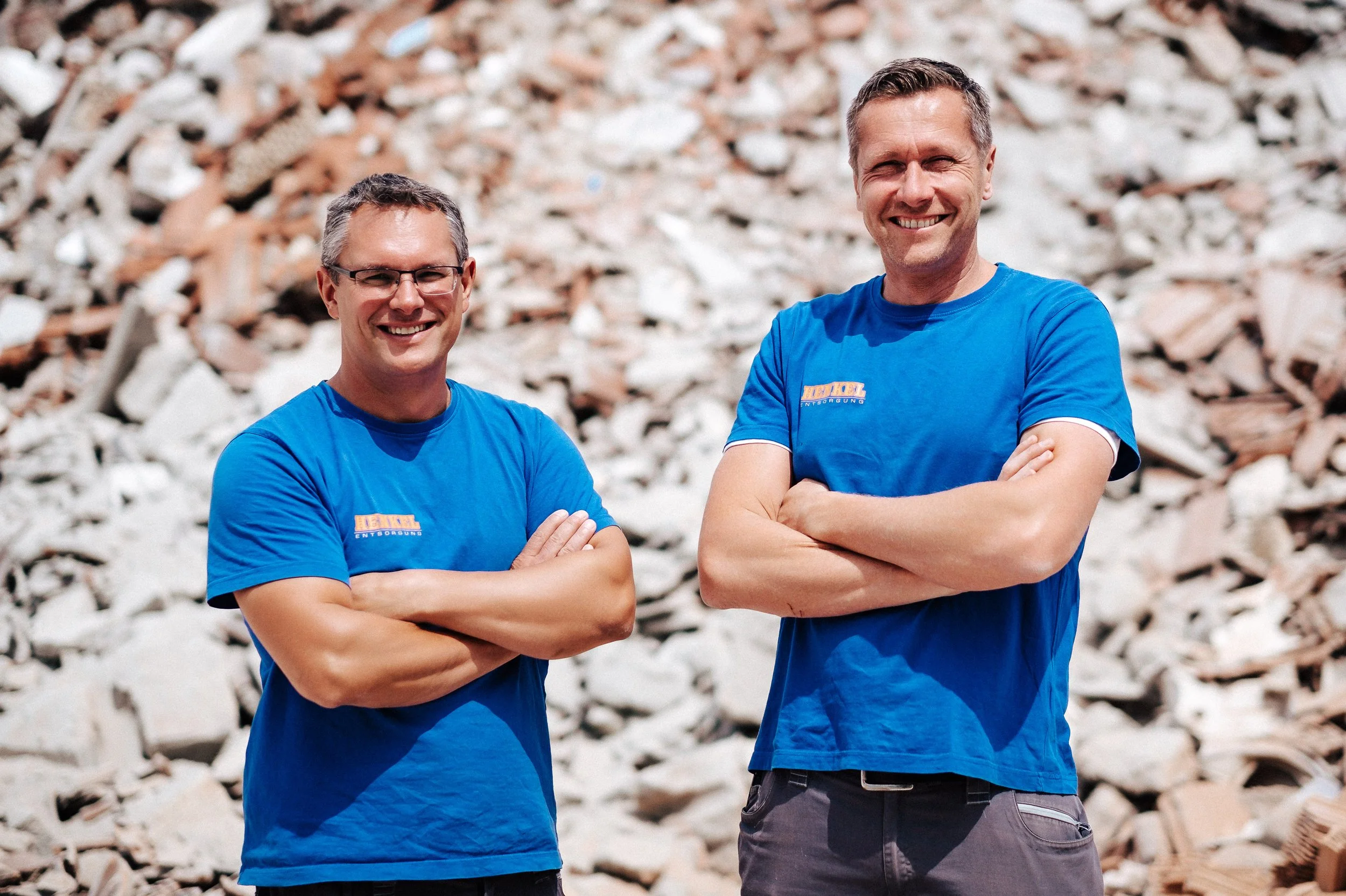 Zwei lachende Männer in blauen T-Shirts stehen vor einem Mauerwerk aus Steinen, beide haben die Arme verschränkt.