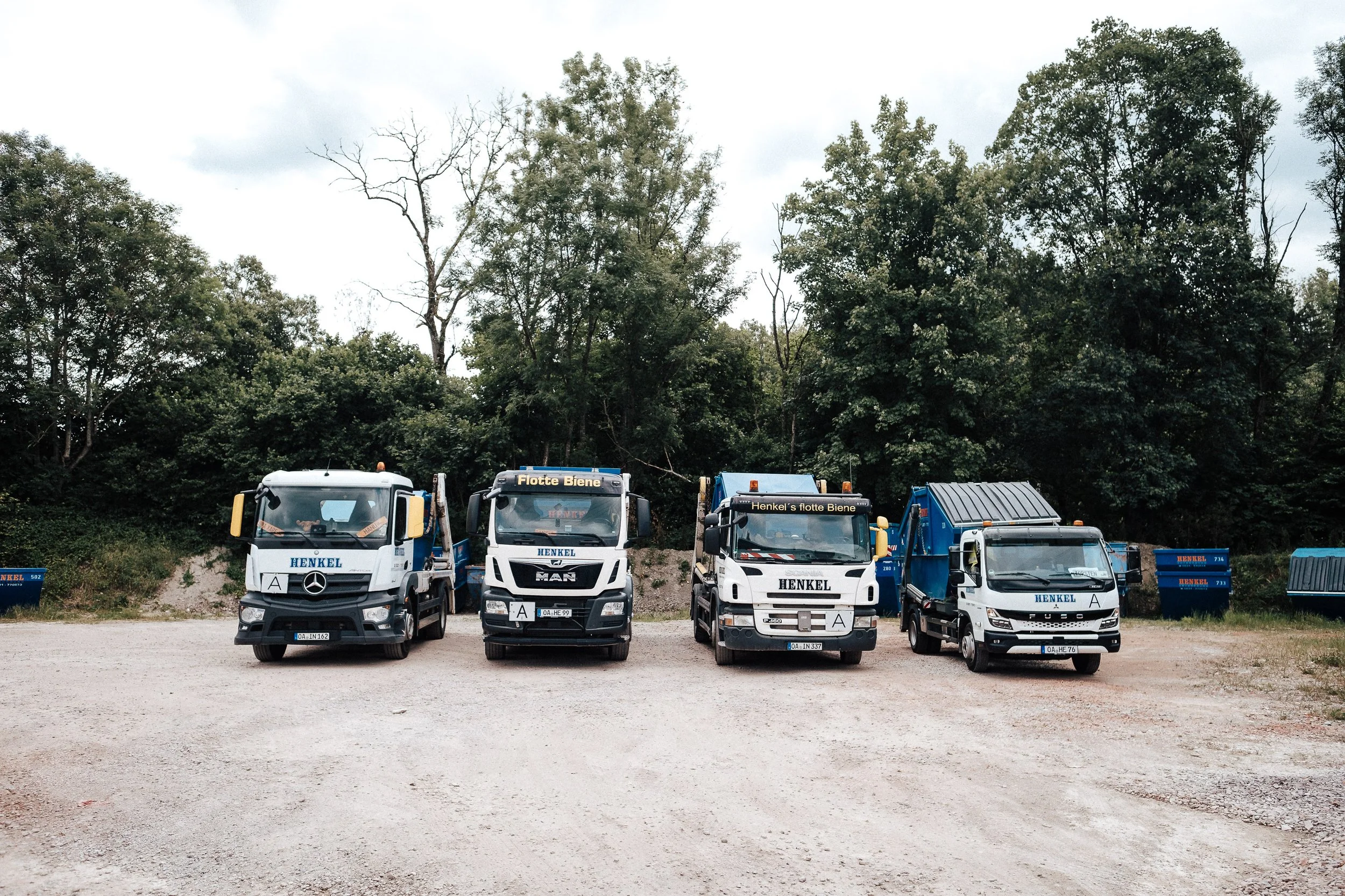 Vier LKWs mit Henkel-Logo parken auf einem unbefestigten Gelände, im Hintergrund Bäume und Container.