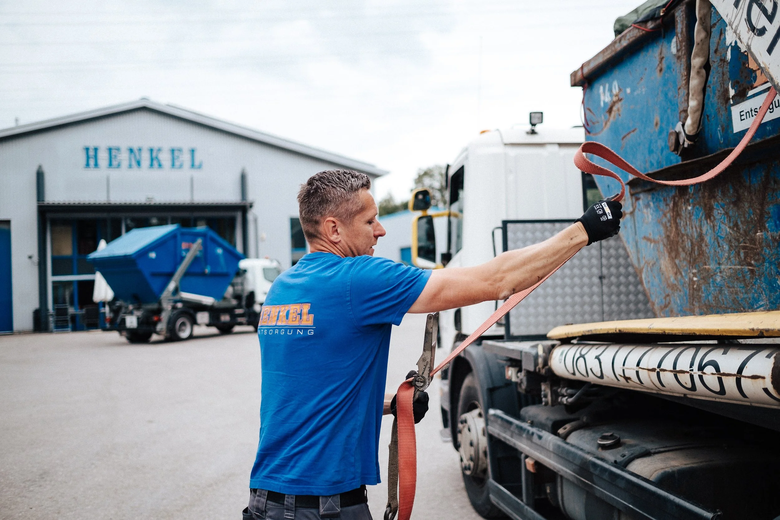 Mann sichert einen Container auf einem Lastwagen mit einem roten Spanngurt vor einem Lagergebäude mit der Aufschrift "HENKEL".