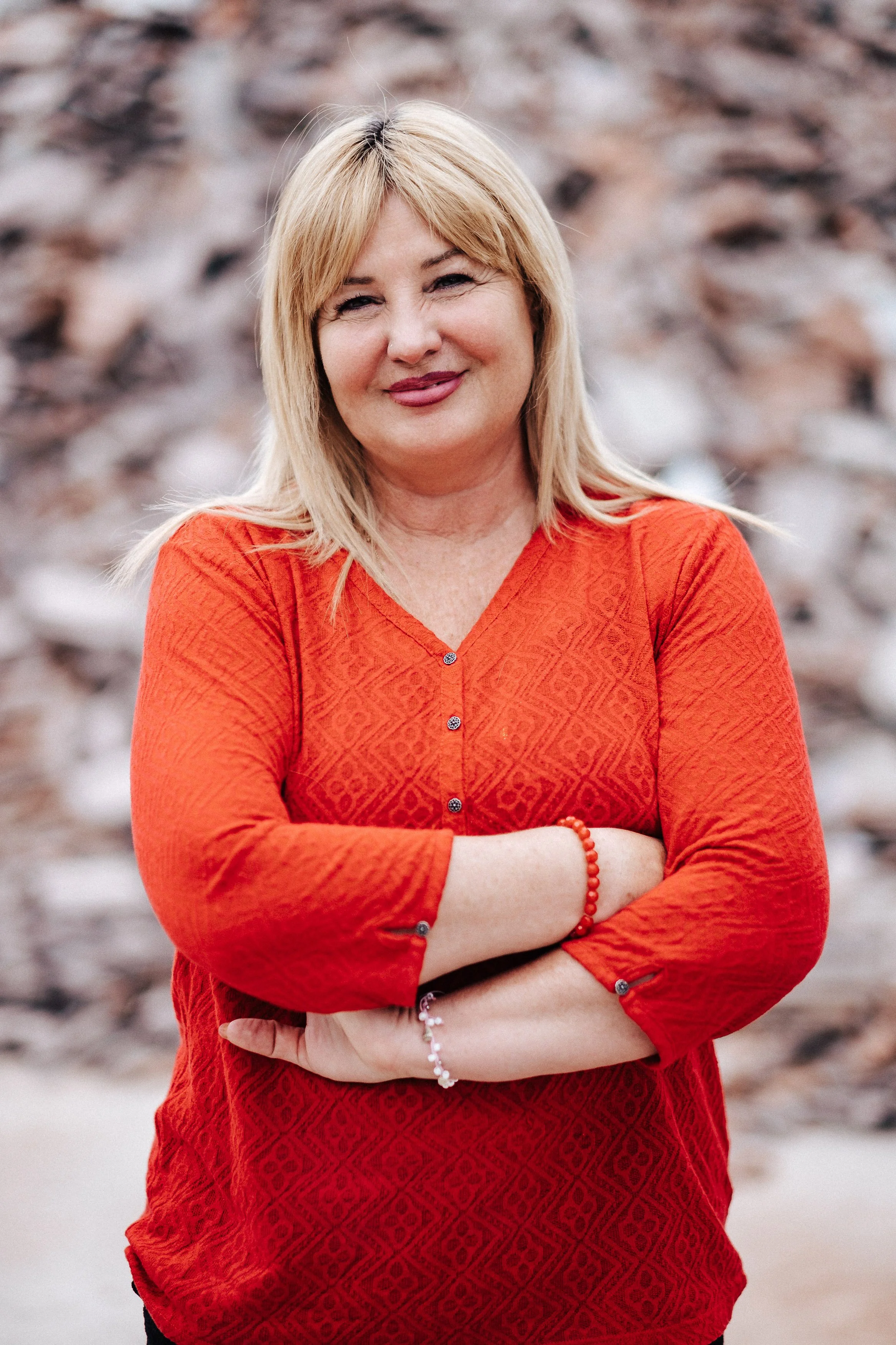 Eine Frau mit blonden Haaren, die ein rotes Oberteil trägt, steht vor einem halb verwitterten Steinhaufen, lächelt und hat die Arme verschränkt.