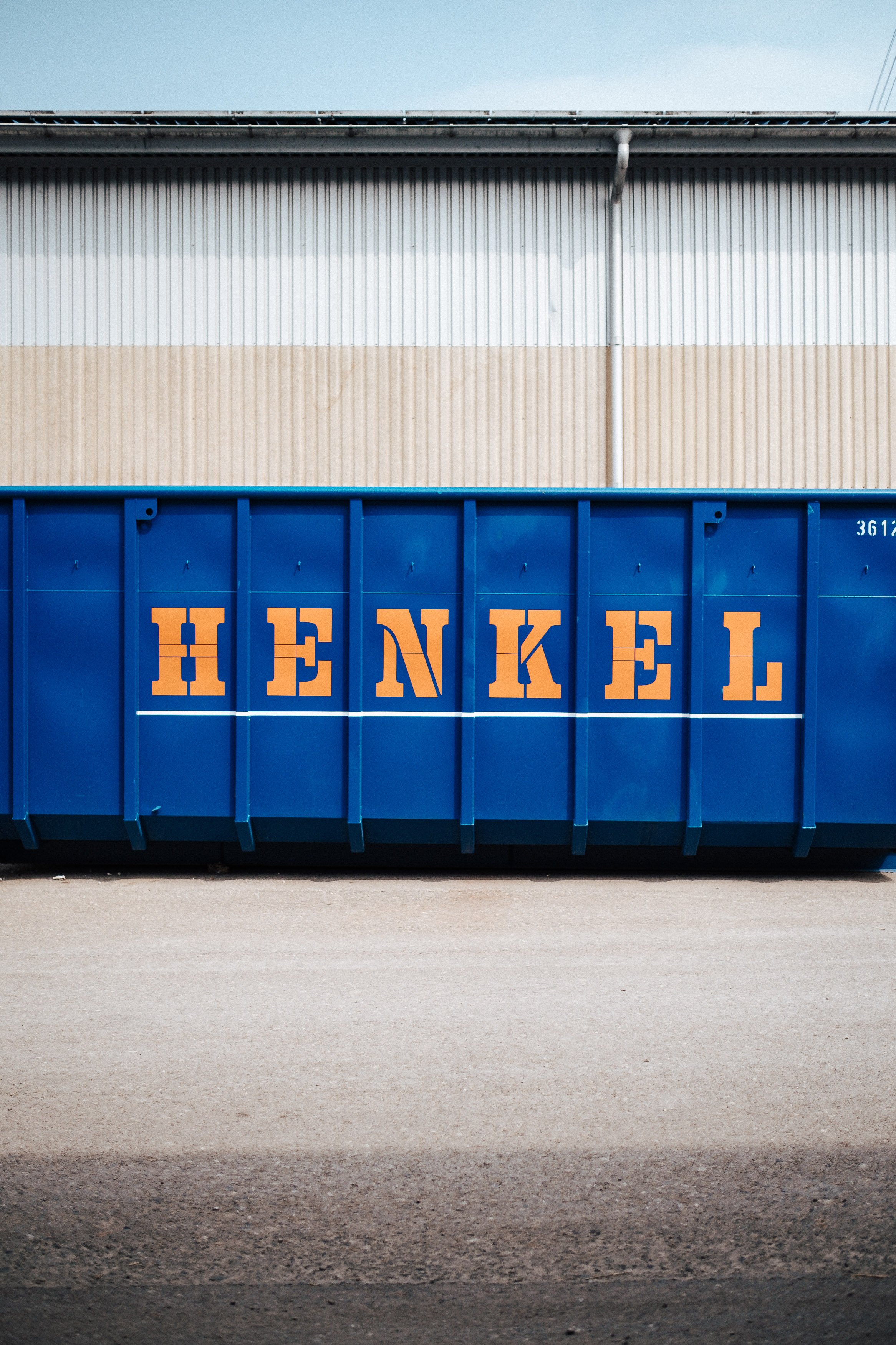 Ein blauer Müllcontainer mit dem Schriftzug 'HENKEL' in orangenen Buchstaben steht vor einer Industriehalle.