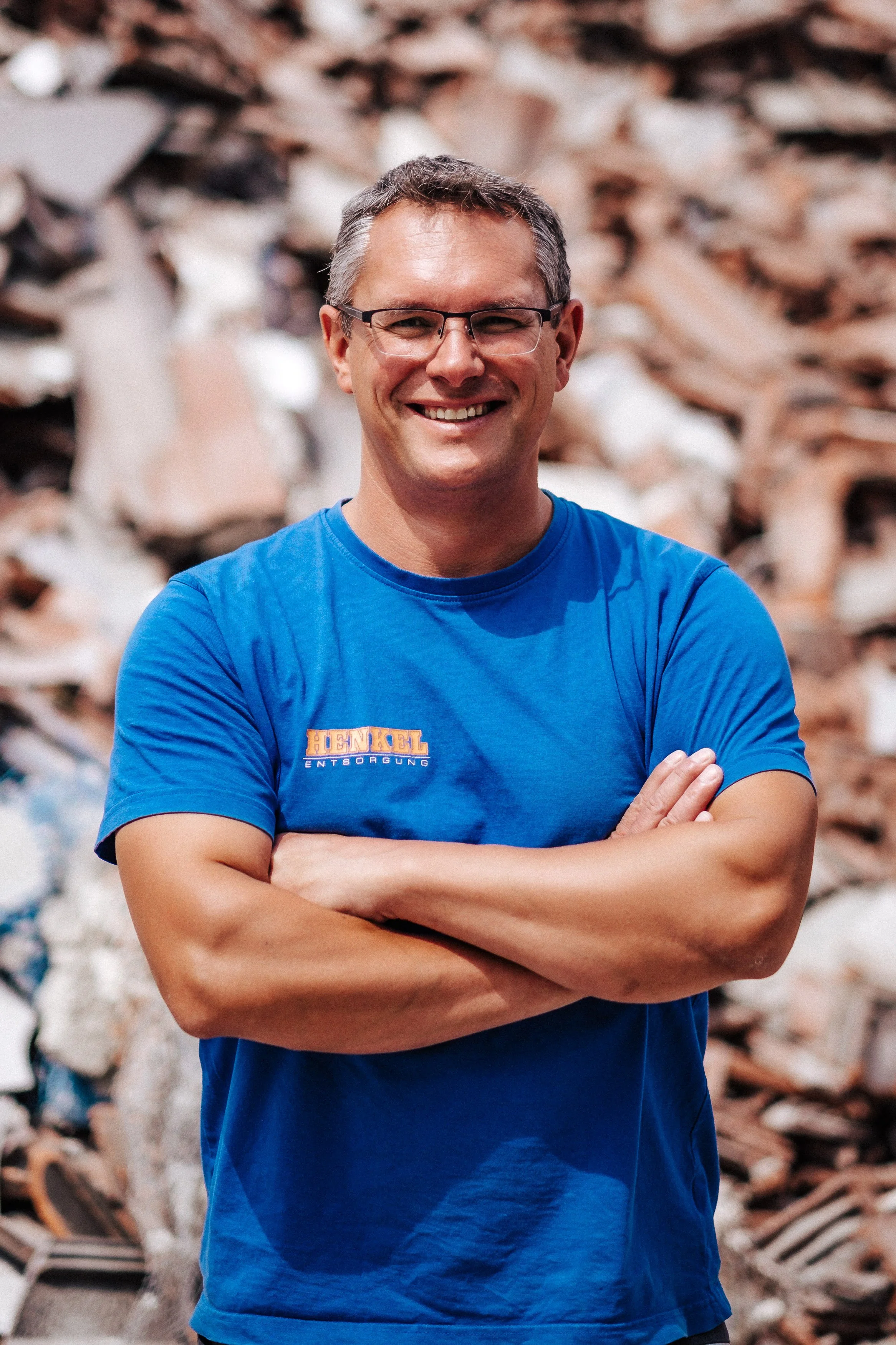 Ein lachender Mann mit Brille in einem blauen T-Shirt steht vor einer Hintergrundwand aus Papier- und Kartonresten, die auf einem Müllhaufen liegen.