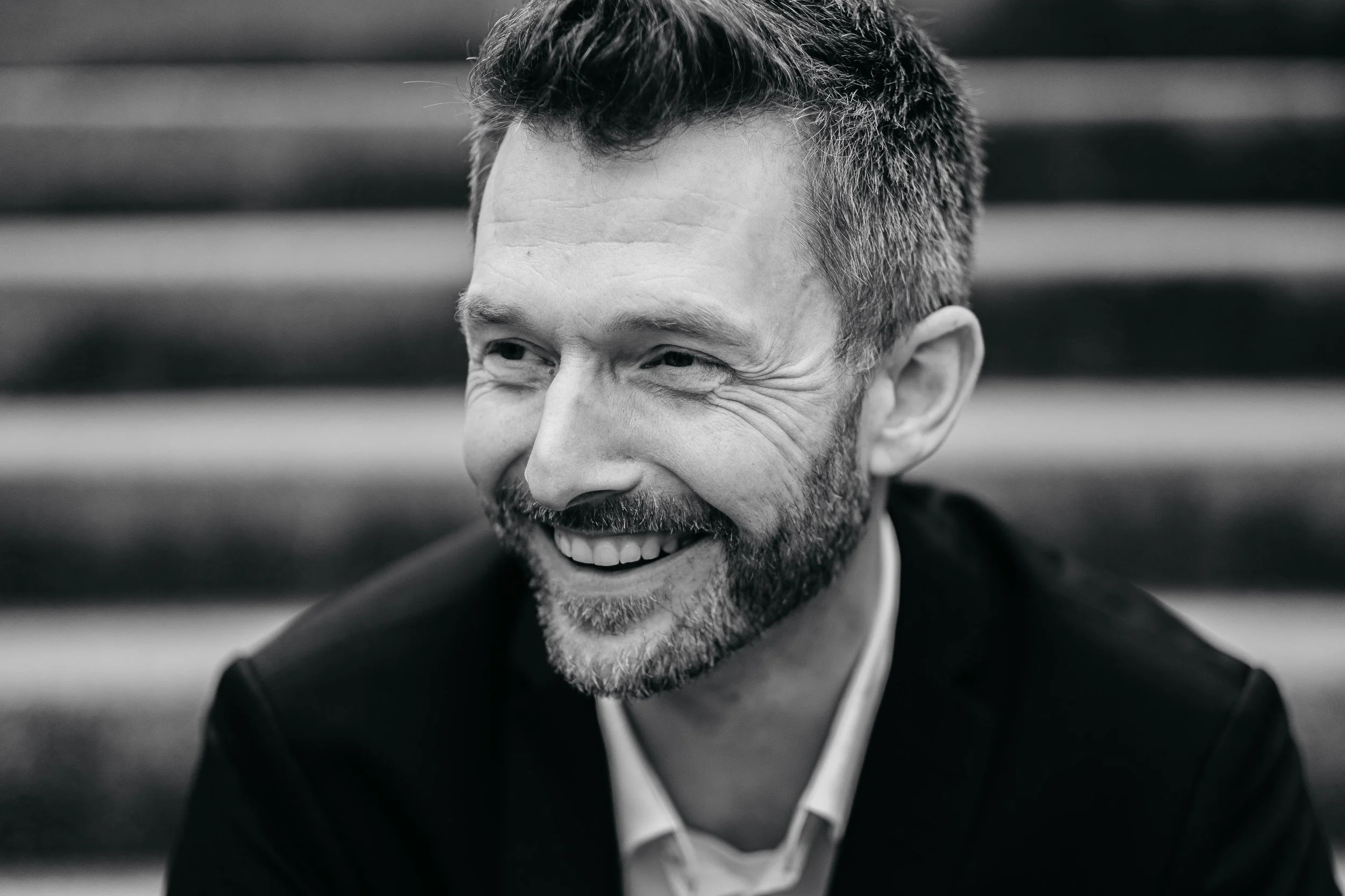 Schwarz-Weiß-Foto eines lächelnden Mannes mit kurzen, dunklen Haaren und Bart, der nach rechts blickt, im Hintergrund Sitzbänke.