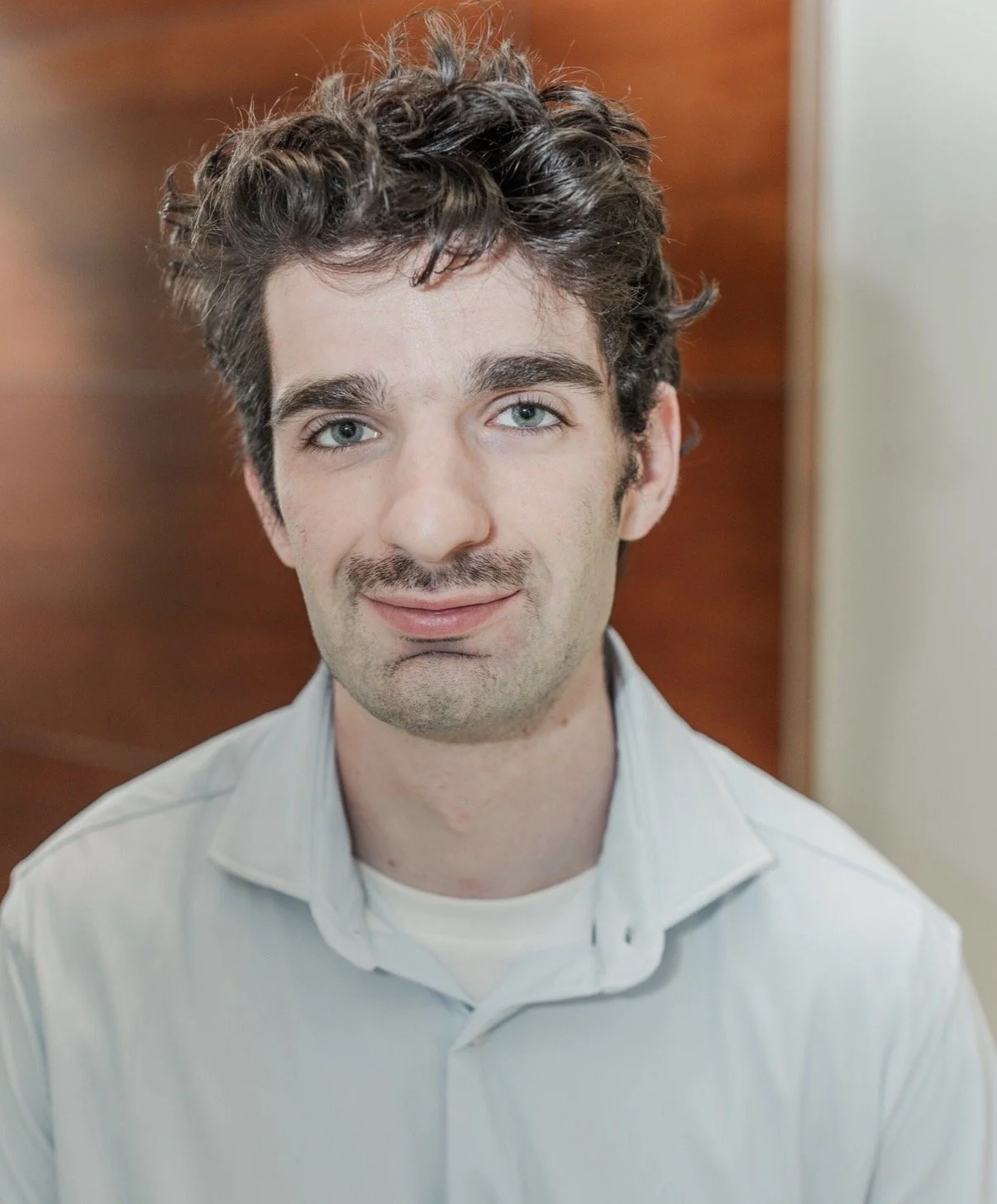 Close-up of a young man with curly brown hair, light blue eyes, and a slight smile, wearing a white shirt, standing indoors in front of a wooden background.