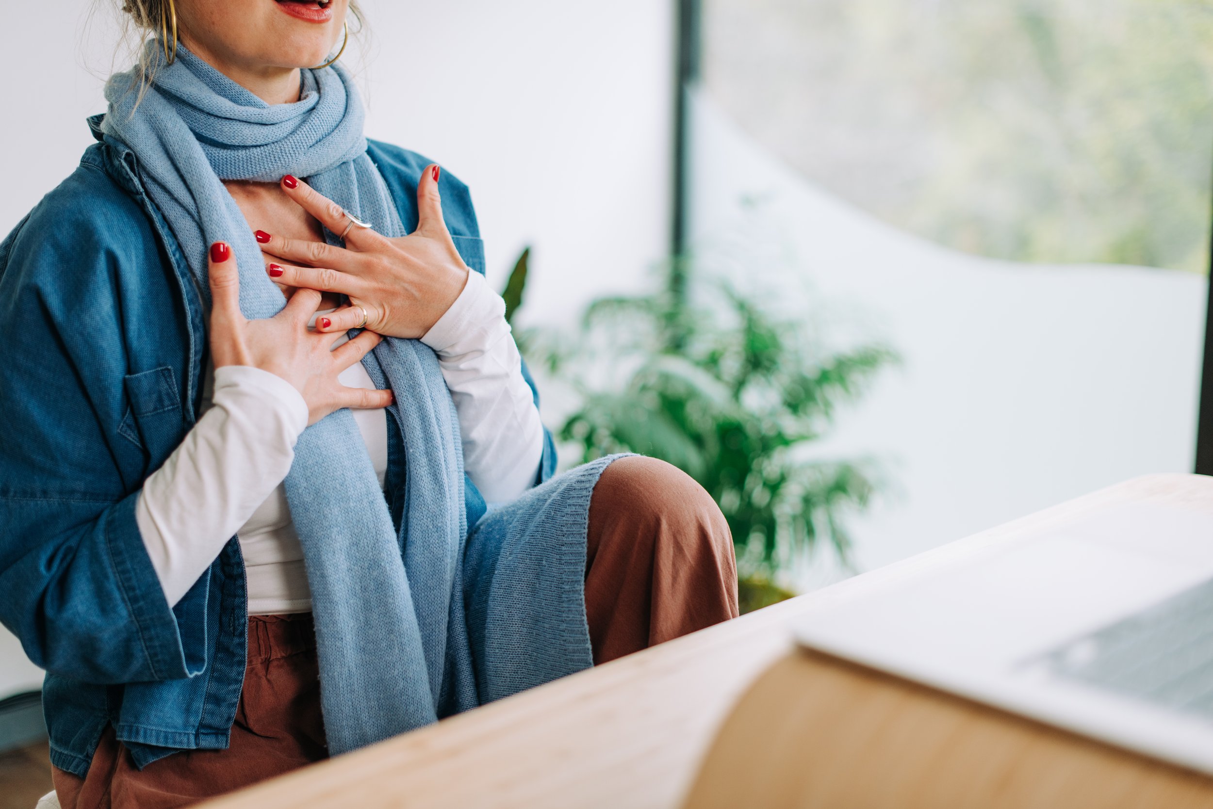 A woman with red lipstick and red nail polish, wearing a light blue scarf and denim jacket, is placing her hands on her chest, appearing to be emotional or grateful.