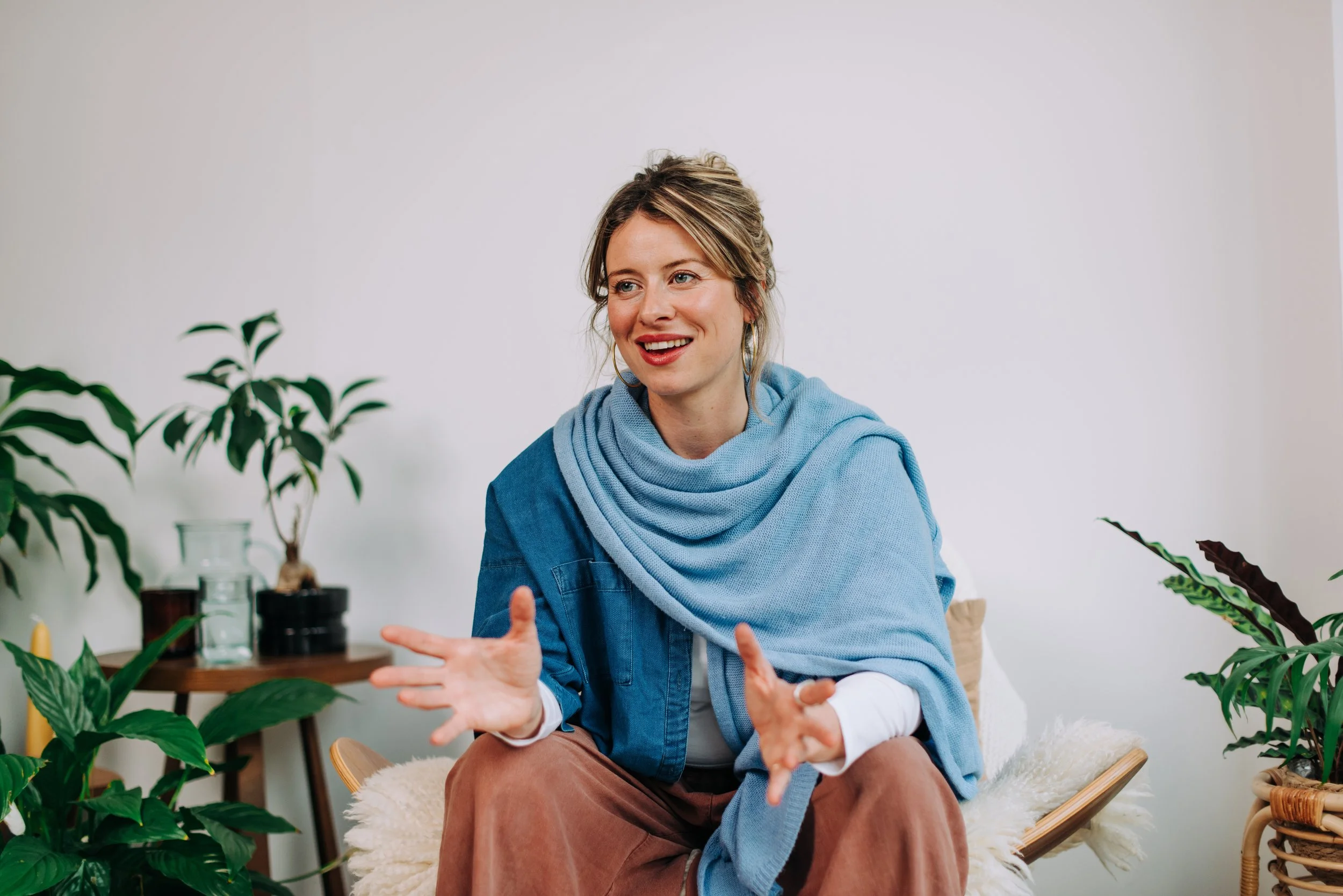 A woman with blonde hair, wearing a blue scarf and jacket, sitting on a chair with a cream fluffy cover, surrounded by green plants, appears to be engaged in a conversation or discussion.