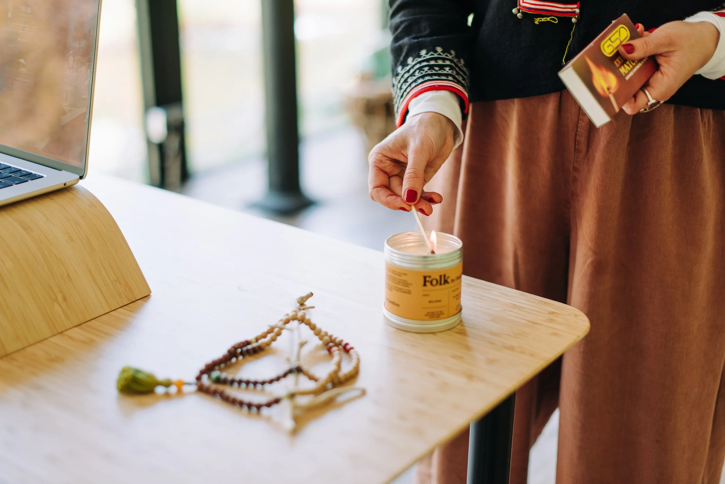 A person lighting a candle with a matchstick in a room with wooden furniture. The person is wearing a dark sweater with colorful embroidery on the sleeve and a brown skirt. There are prayer beads and a laptop on the table, and a matchbox in their hand.