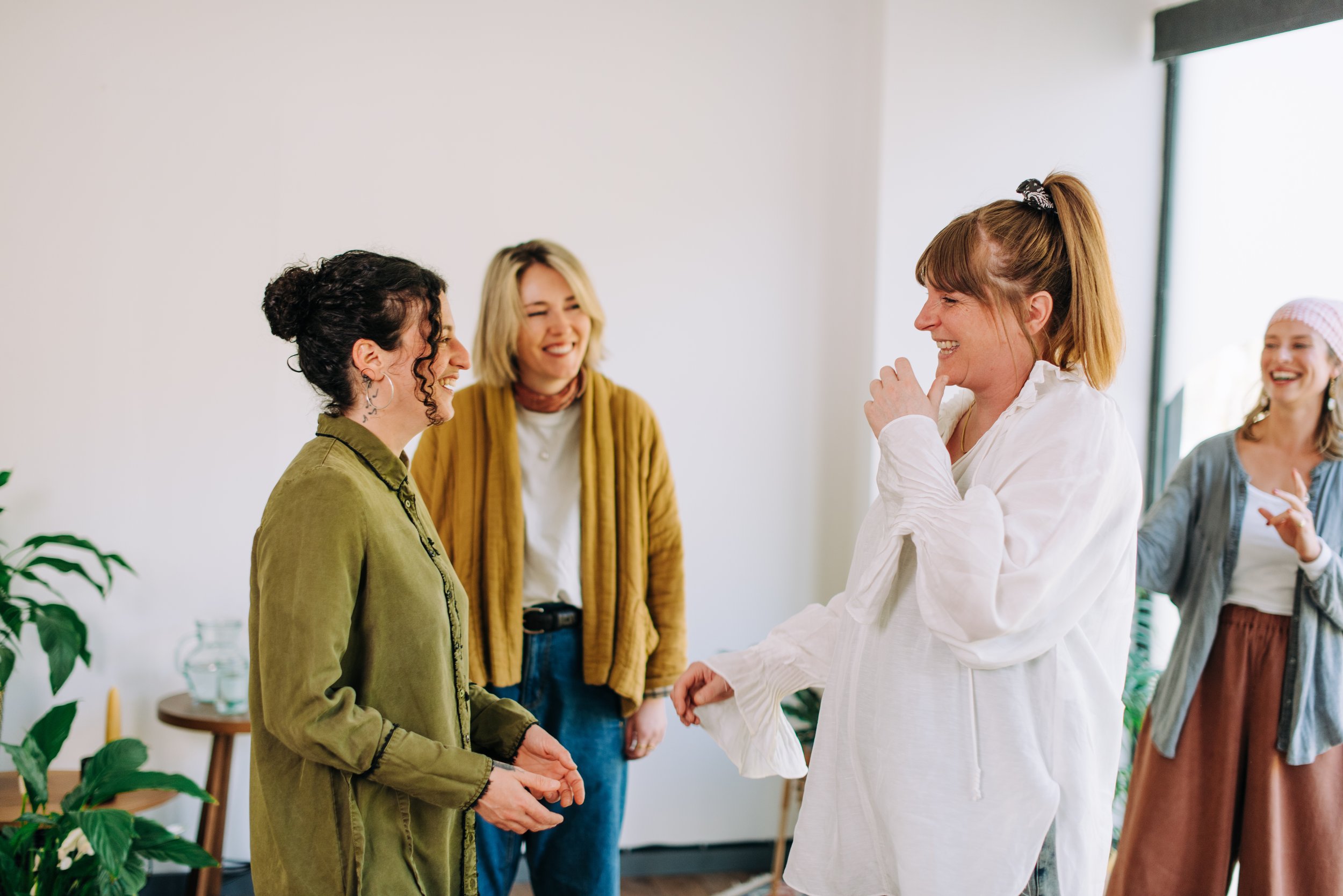 Four women are engaged in a lively conversation indoors, smiling and laughing. They are casually dressed, and the atmosphere appears joyful and friendly.