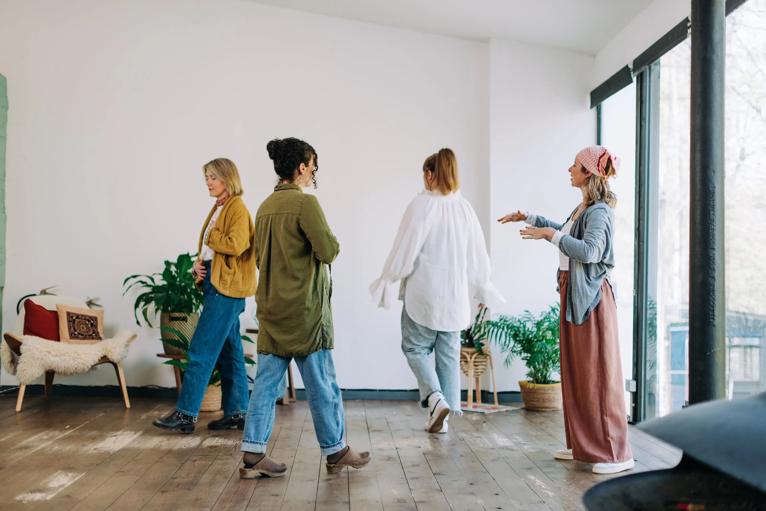 Four women socialize in a bright, modern room with large windows and indoor plants, engaged in conversation or activity.