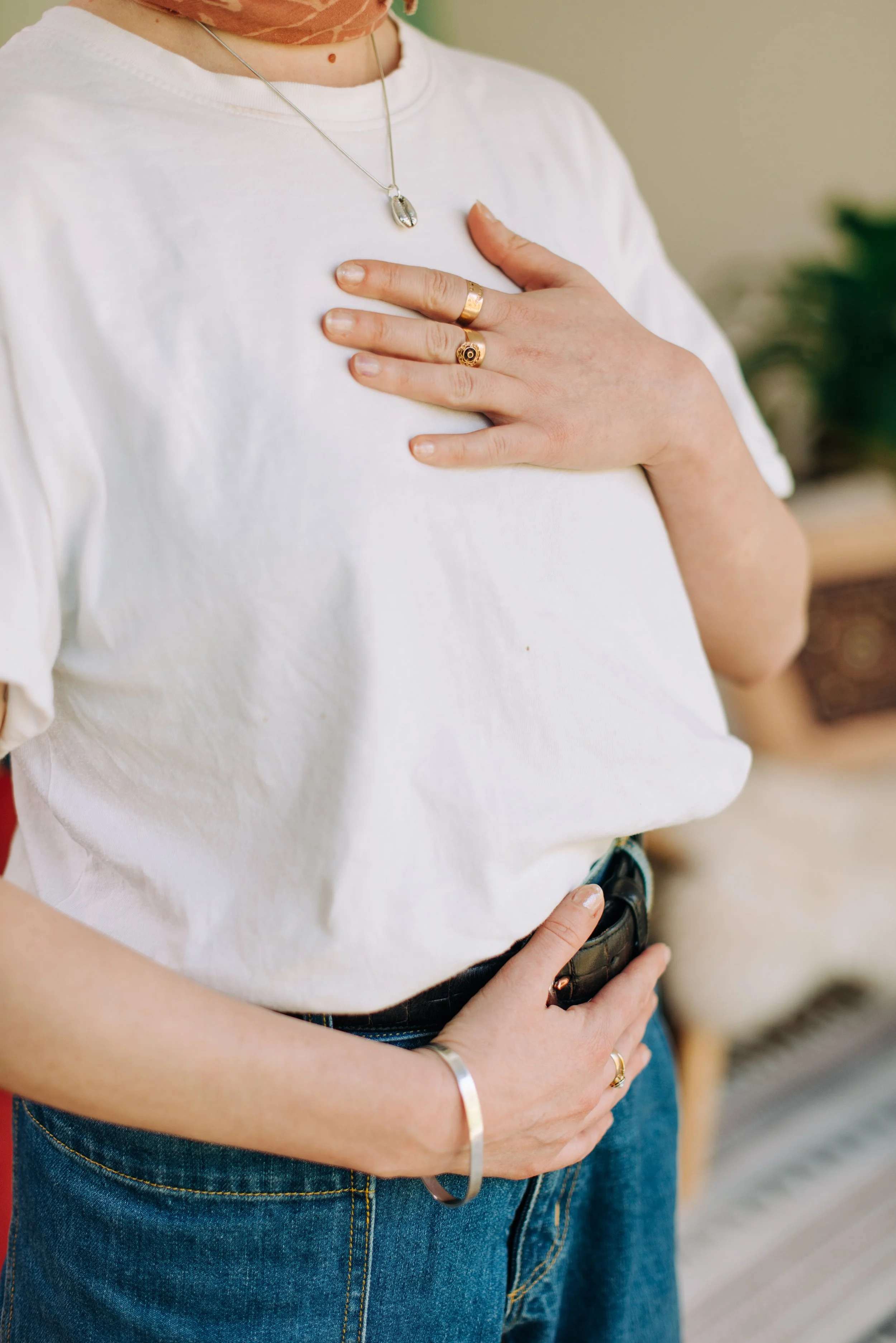 Close-up of a person wearing a white t-shirt, multiple rings, a necklace, and a bracelet, with one hand on their chest and the other resting on their waist, holding a phone.