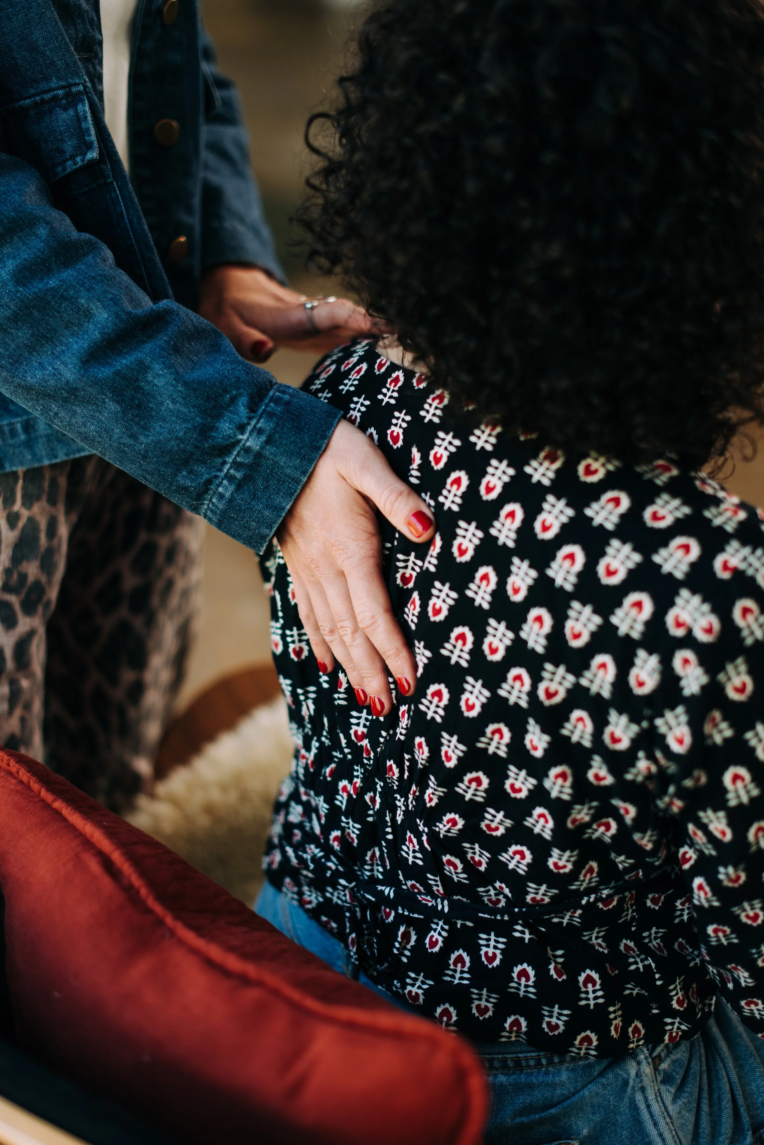 Close-up of two people holding hands, one wearing a denim jacket and the other in a top with a red, white, and black pattern, sitting in a cozy setting.