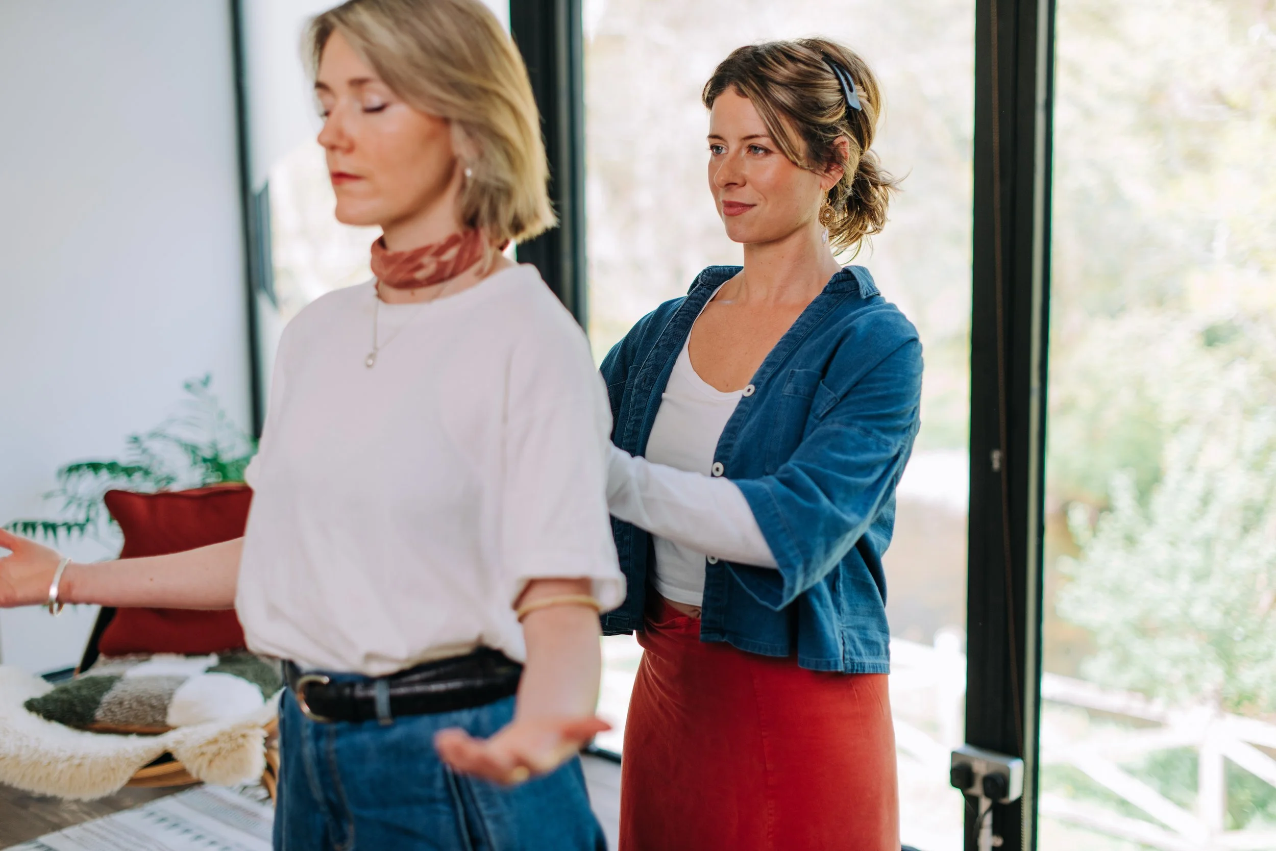 Two women standing indoors near large windows, one woman is in the foreground holding a tray and the other woman is in the background wearing headphones, looking at the woman with the tray.