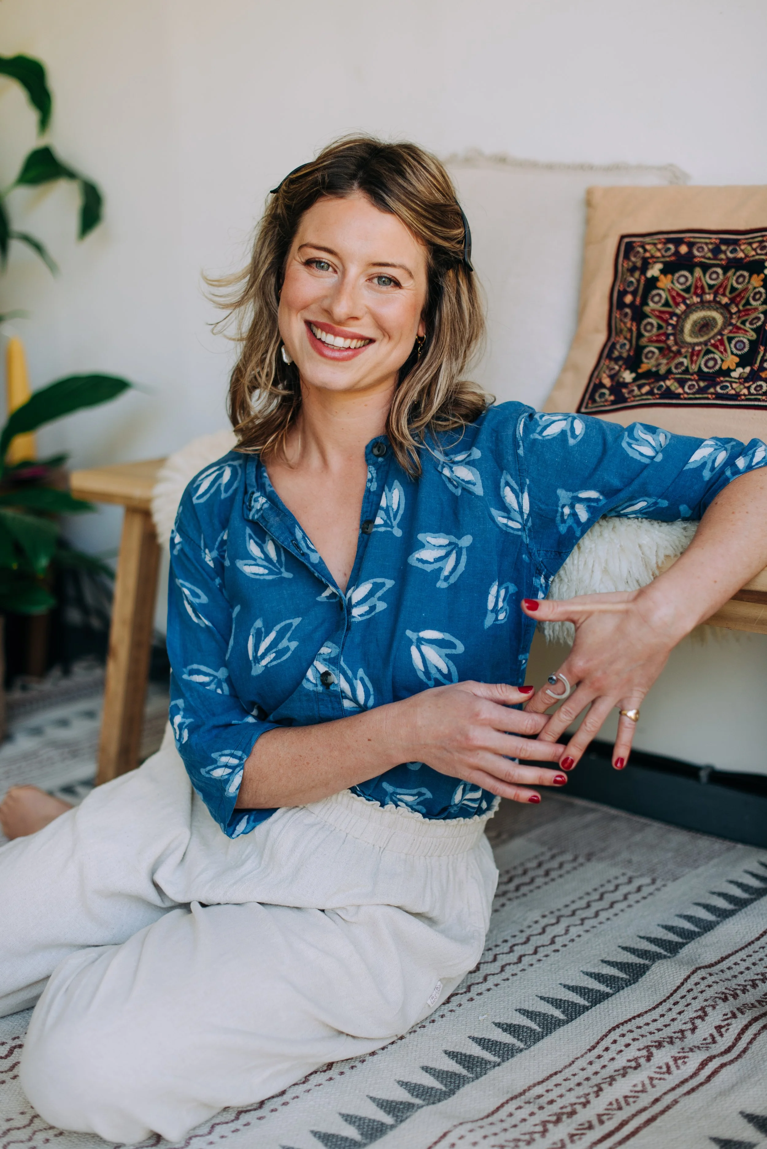 A smiling woman with shoulder-length wavy hair, wearing a blue patterned blouse and white pants, sitting on a patterned rug in a cozy indoor space with cushions and a potted plant in the background.