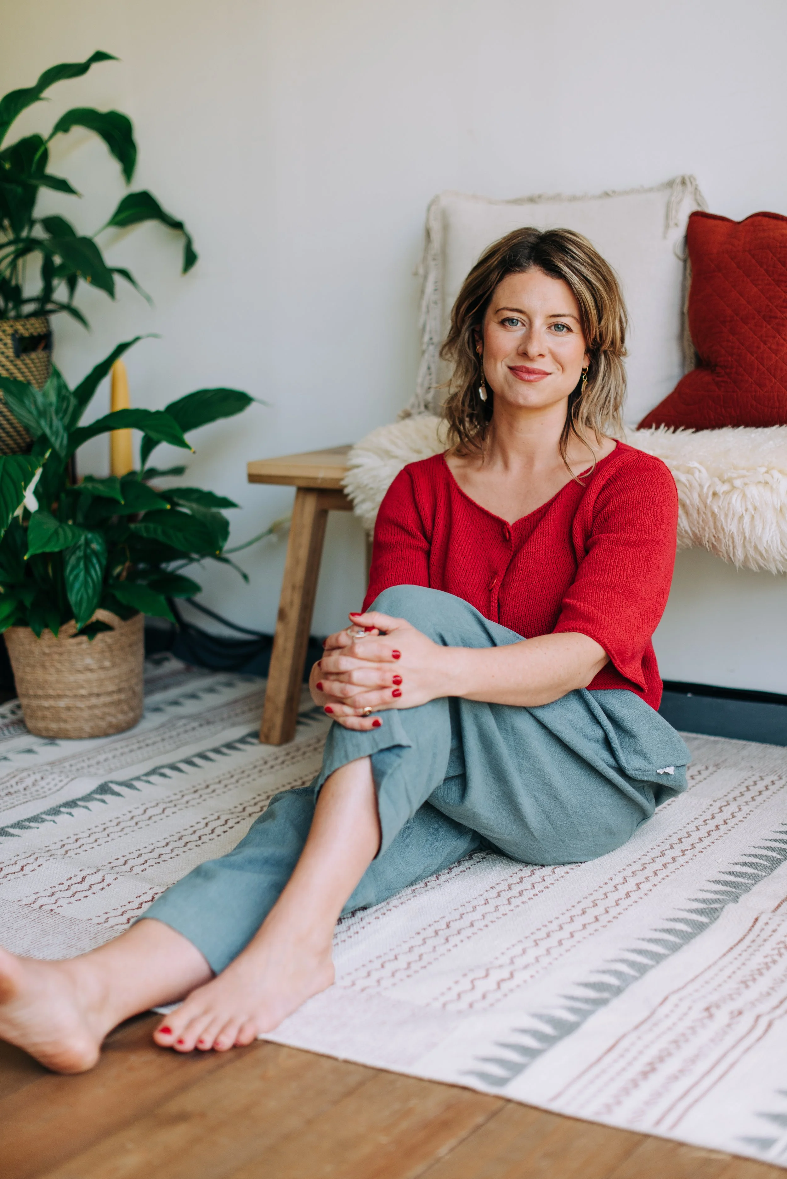 A woman sitting on a patterned rug on the floor with her legs crossed, wearing a red sweater and gray pants, with a smile, in a room with a green plant, a wooden side table, and red and white pillows.