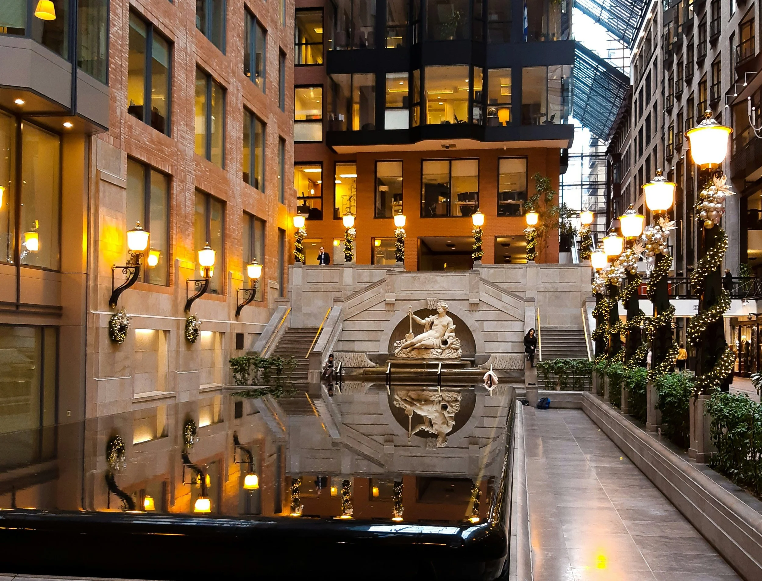 Un intérieur d'un centre commercial ou un bâtiment avec une décoration festive, comprenant des guirlandes de Noël, de nombreuses lampes, une fontaine avec une statue et un escalier en pierre, vue depuis l'intérieur, vue partielle d'une voiture en premier plan.