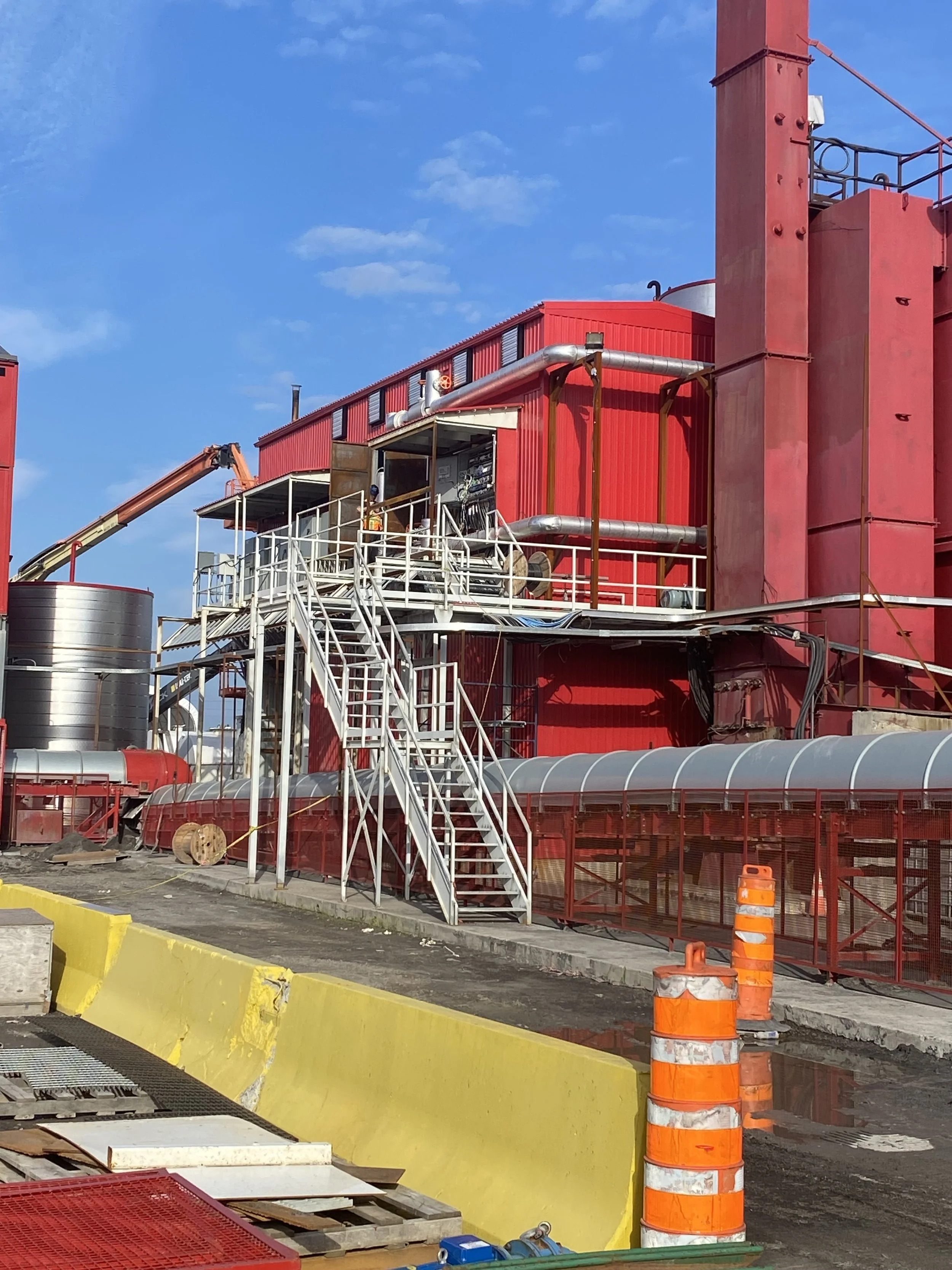 Construction d'une structure industrielle rouge avec des tuyaux en acier, des escaliers en métal, et des barrières de sécurité orange, sous un ciel bleu.