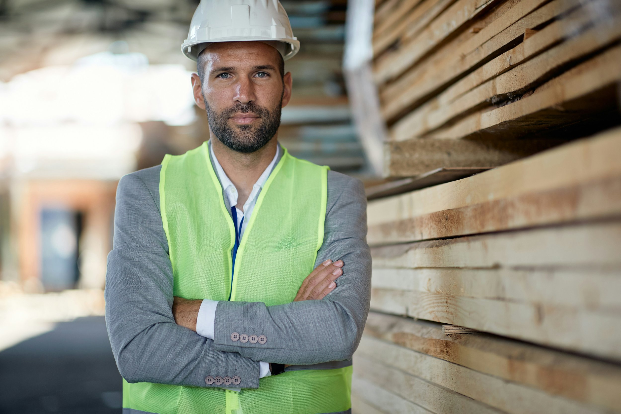 Un homme portant un casque de sécurité et un gilet de sécurité vert foncé regarde d'un air sérieux, avec des bras croisés, devant un fond de planches en bois dans une zone de stockage ou un chantier.