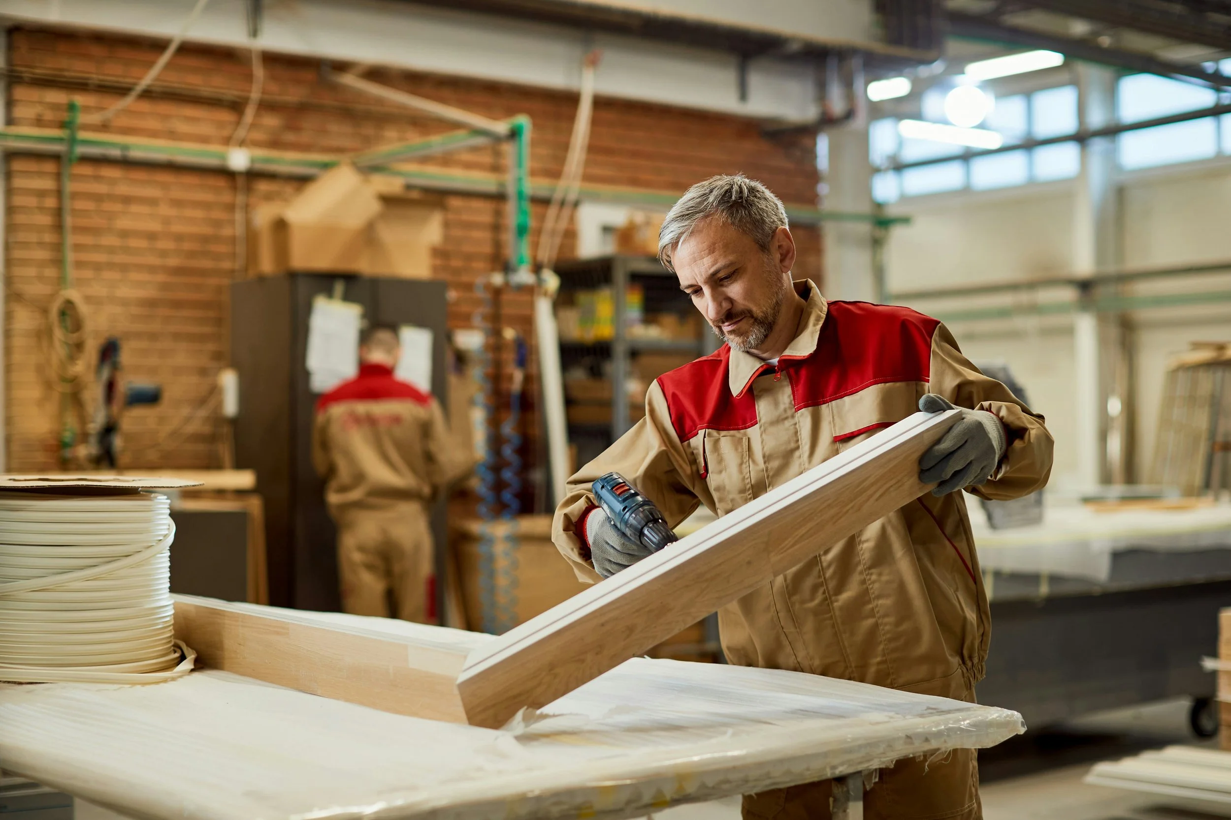 Un homme en uniforme de travail, dans un atelier de menuiserie, utilise une perceuse sur un morceau de bois. Deux autres personnes travaillent en arrière-plan. L'environnement inclut des murs en briques et de grandes fenêtres.