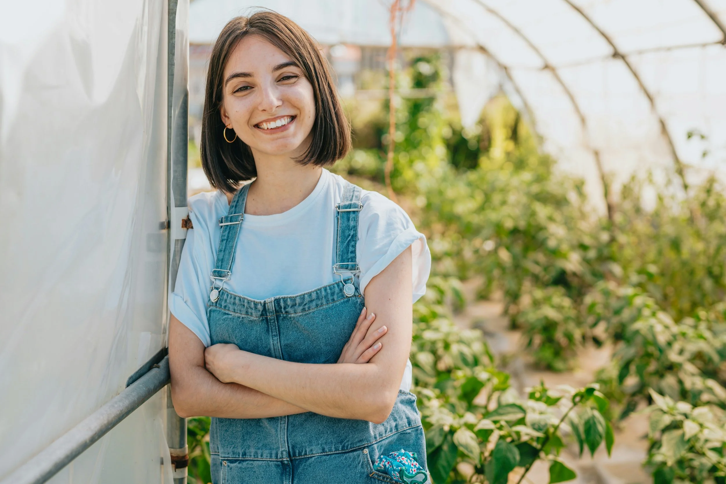 Jeune femme brune avec un sourire, portant un t-shirt blanc et un jean avec salopette, debout dans une serre ou un jardin avec des plantes vertes en arrière-plan.