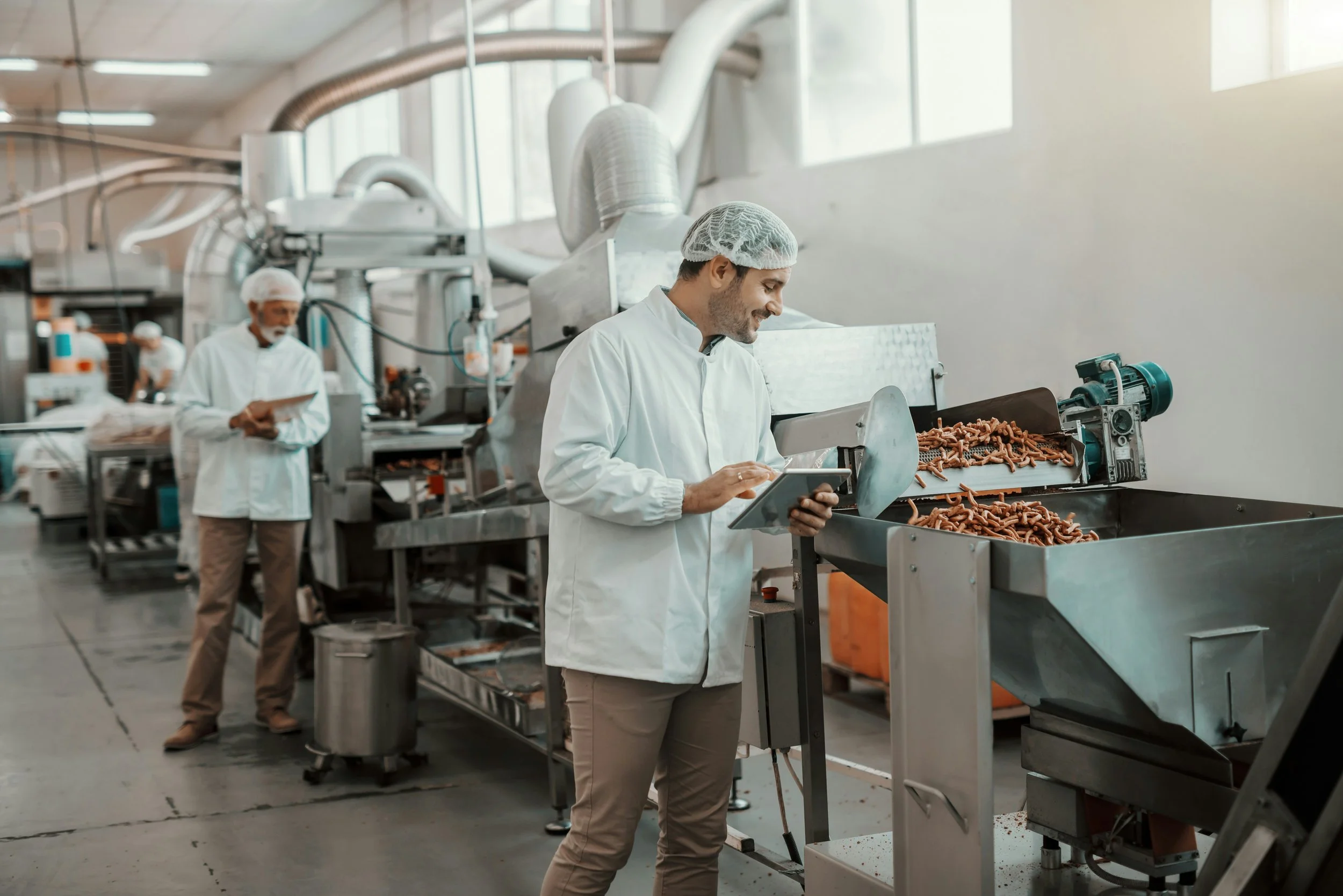 Travail dans une usine alimentaire avec plusieurs employés en uniforme et charlottes, opérant une machine pour traiter de la viande.