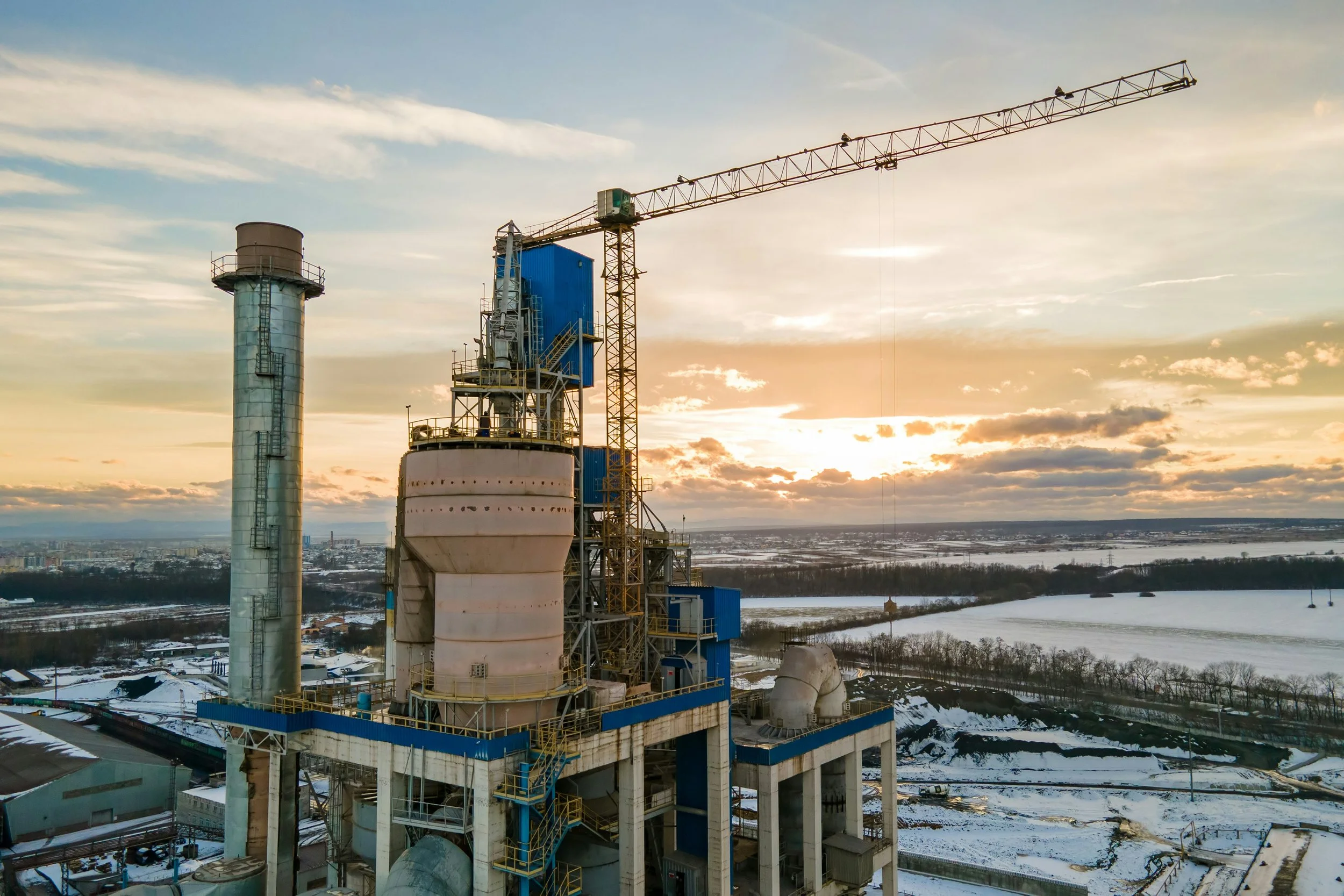 Une centrale électrique en construction avec une tour de refroidissement, une grue et des structures en acier, avec un paysage enneigé au loin, sous un ciel nuageux au coucher du soleil.