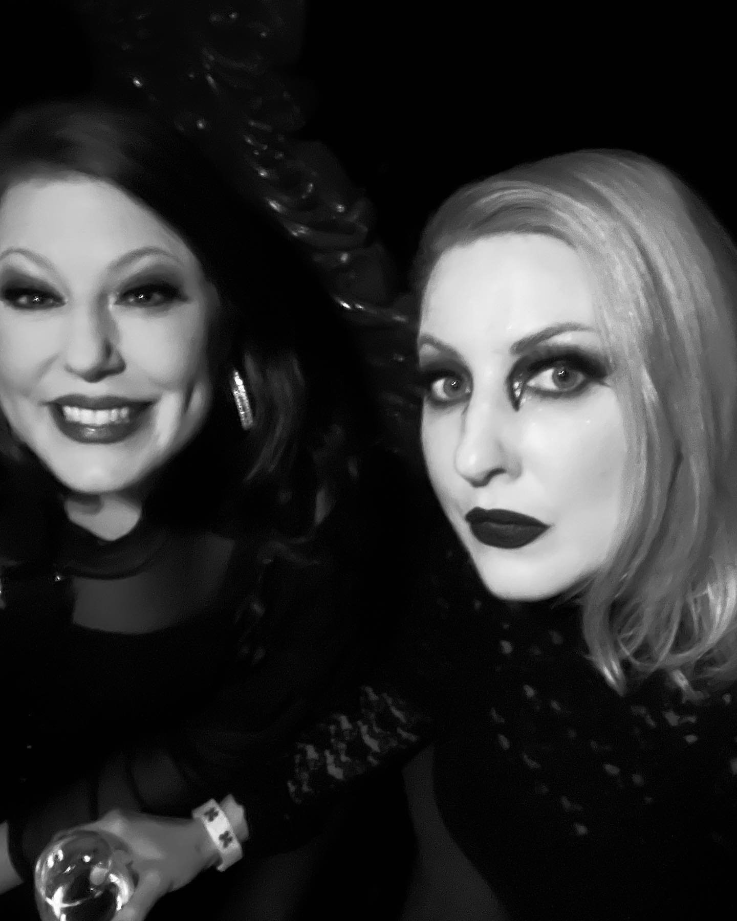 Black and white photo of two women with dramatic makeup, with the woman on the left smiling and the woman on the right with a serious expression, both wearing dark clothing and looking at the camera.