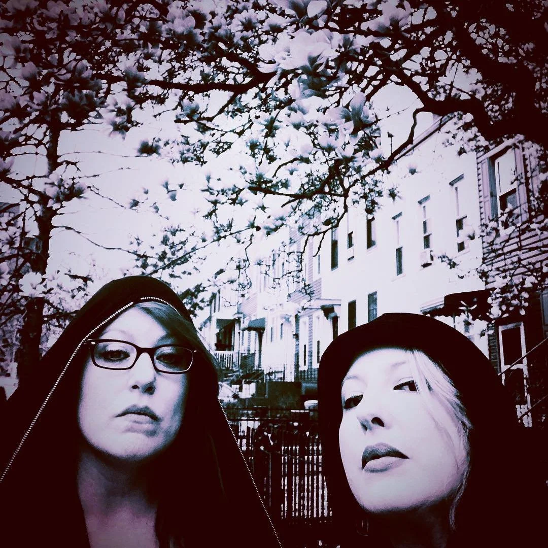 Two women with pale skin and dark hair, one wearing glasses and a hooded jacket, the other with a hat, posing under a blooming tree with white flowers on an urban street.
